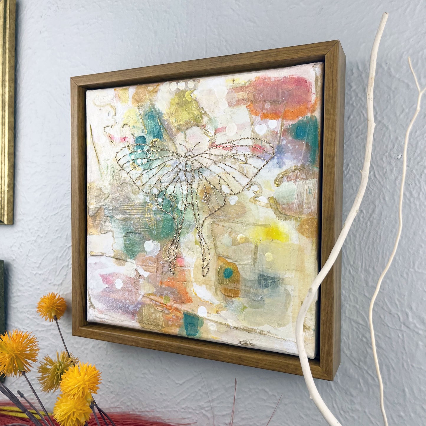 angled view of a colorful square painting in a wood frame, with a line drawing of a luna moth hand stitched through it in metallic gold thread, hanging on a wall