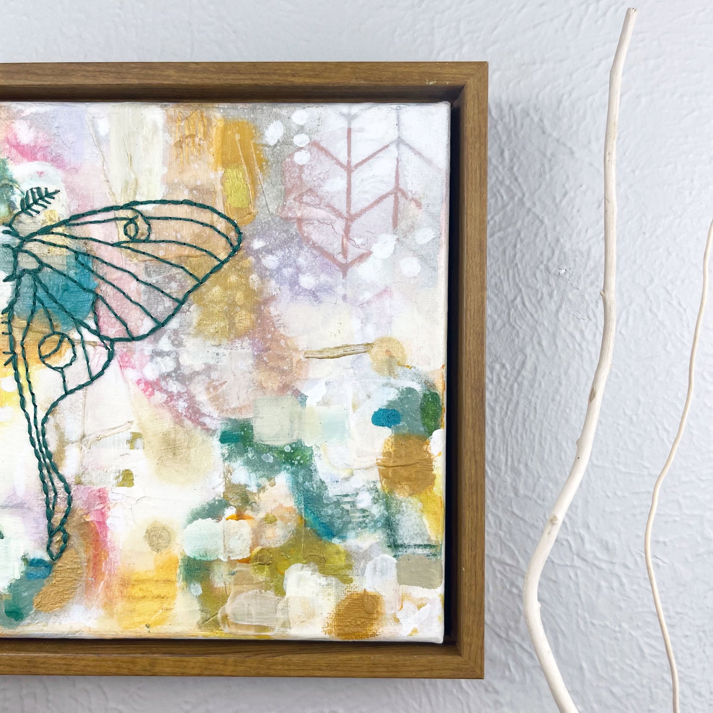 off center view of a colorful square painting with a hand stitched half of a luna moth in emerald green embroidery floss, in a wood frame hanging on a wall