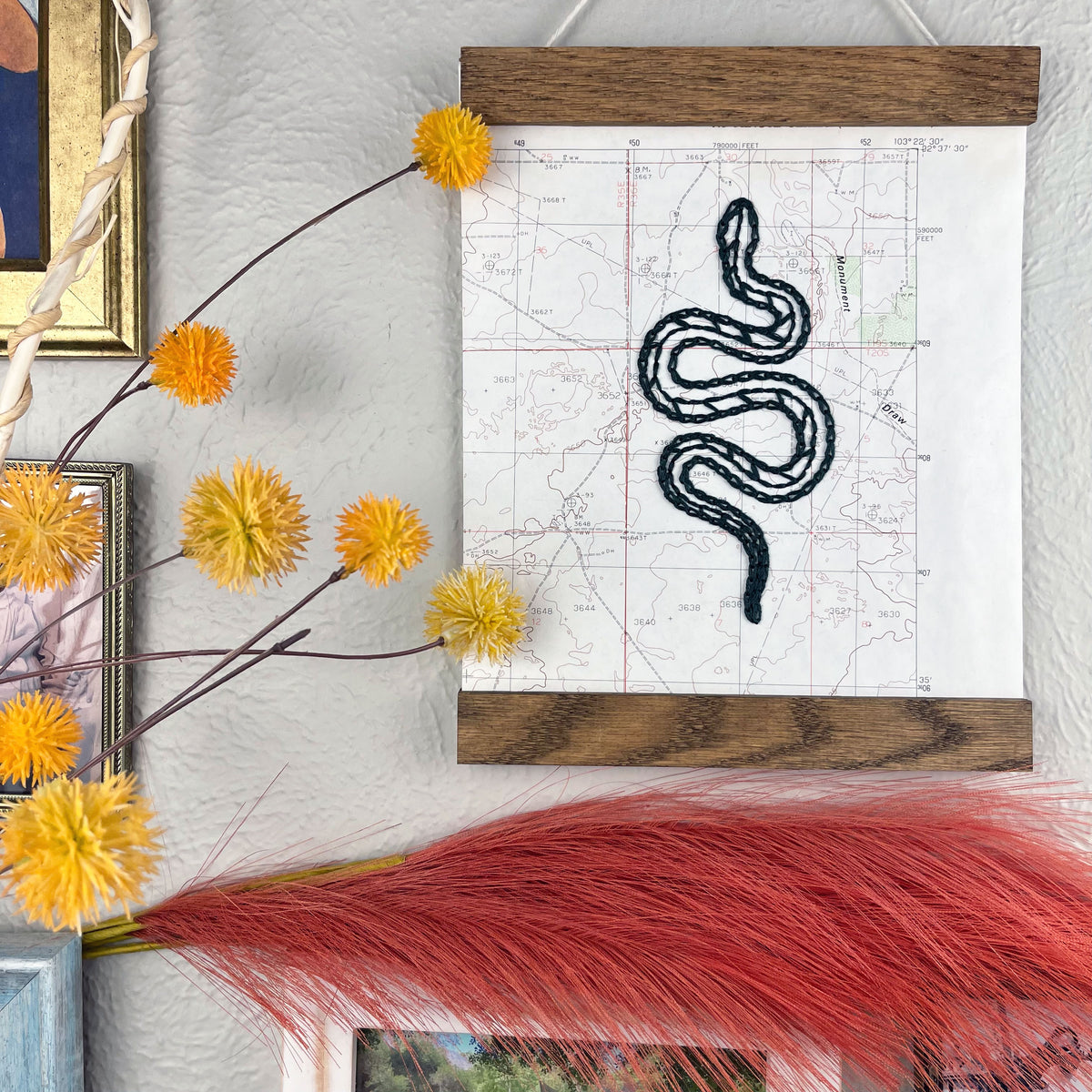 a vintage USGS map of New Mexico hand embroidered with a dark green snake, framed on top and bottom with a rustic wood magnetic frame hanging on a grey wall with framed art and botanical decorations around it