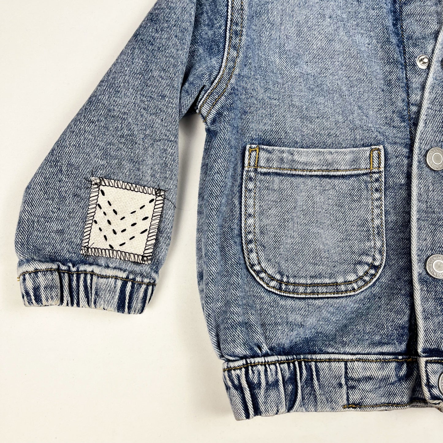 close up view of wearer's right half of a denim jacket, with a small natural colored square patch on the sleeve near the cuff, stitched with three black chevron lines, overlocked in black thread