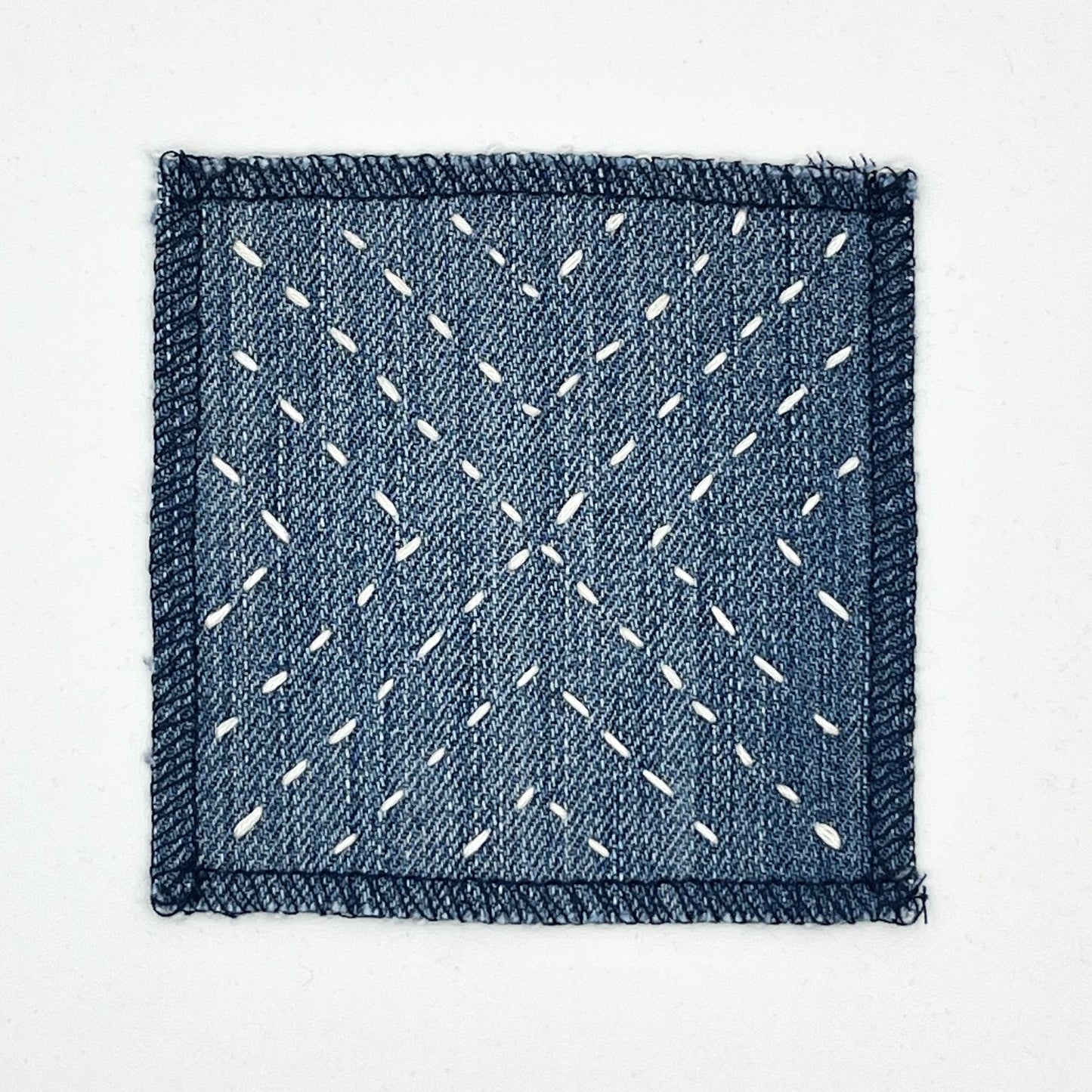 a square patch made out of denim, handstitched with ivory running stitches in a radiating X pattern, with overlocked edges, on a white background