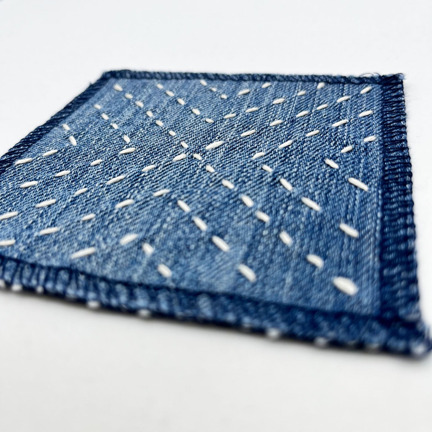 close up angled view of a square patch made out of denim, handstitched with ivory running stitches in a radiating X pattern, with overlocked edges, on a white background