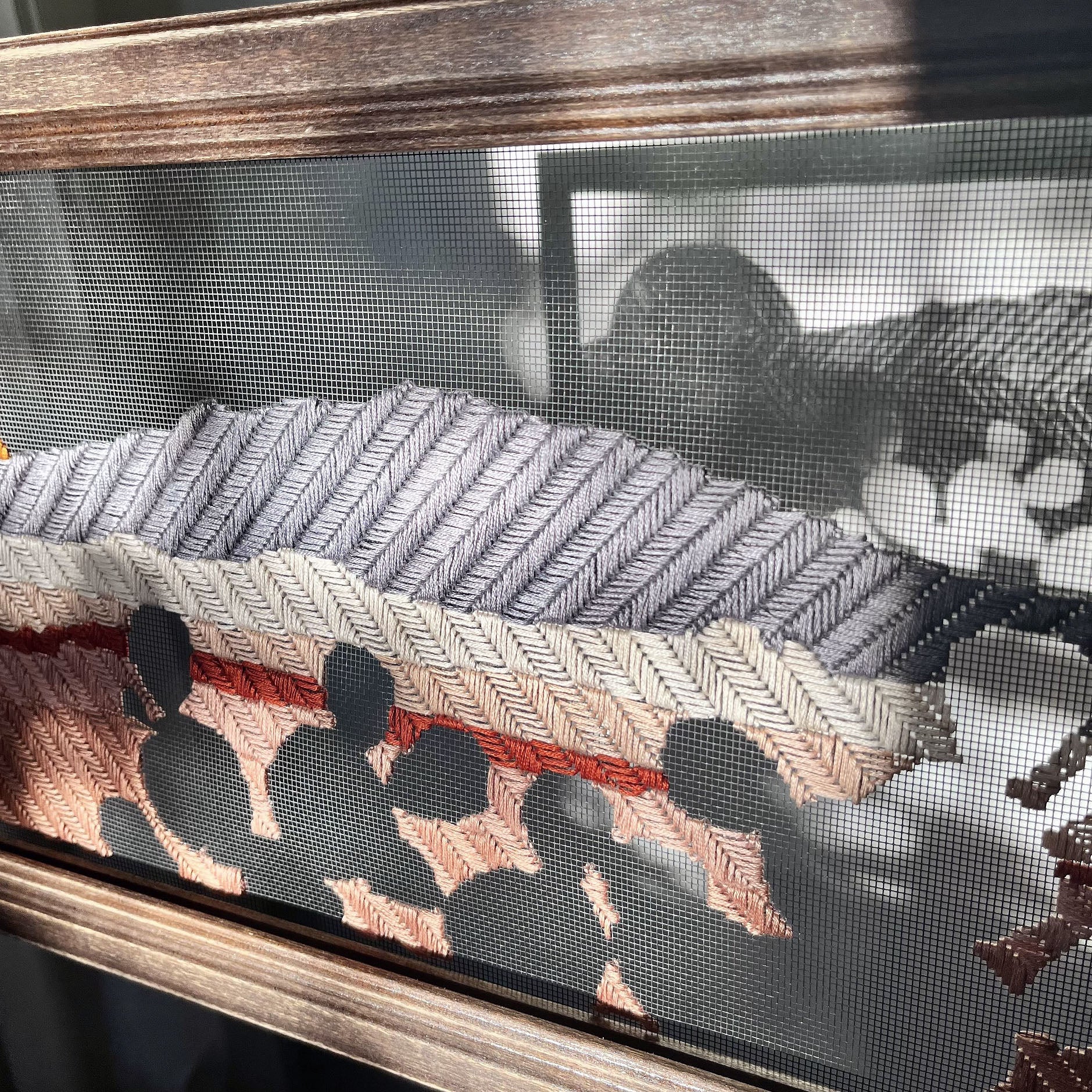framed window screen hand stitched with the image of a mountain, volcanoes and sun, with negative space prickly pear cactus in the foreground, and the shadow of the stitches showing through the screen on the wall behind it