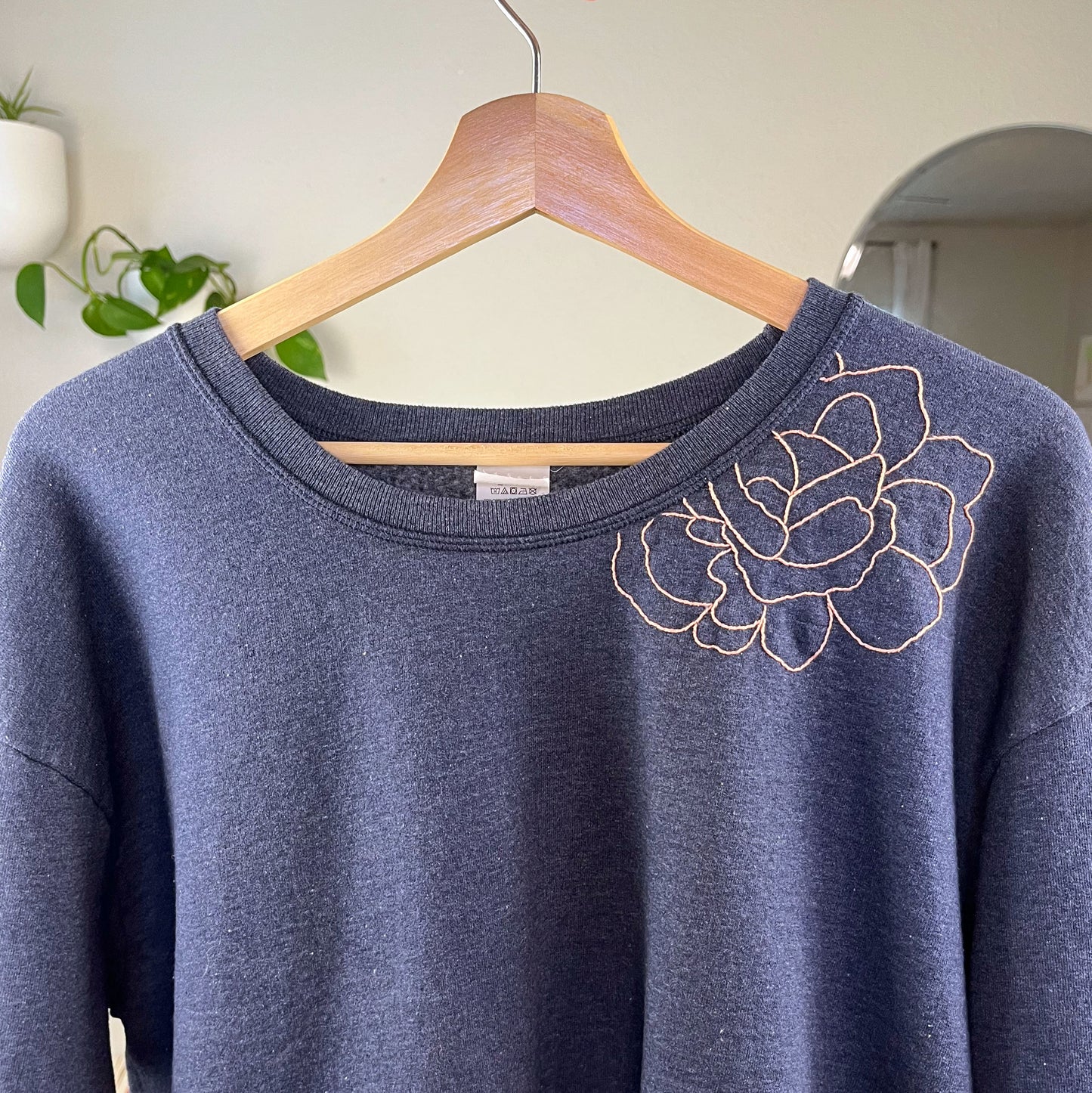 a navy colored sweatshirt with a peach hand embroidered outline of a peony on the front shoulder and into the front neckline, on a wood hanger