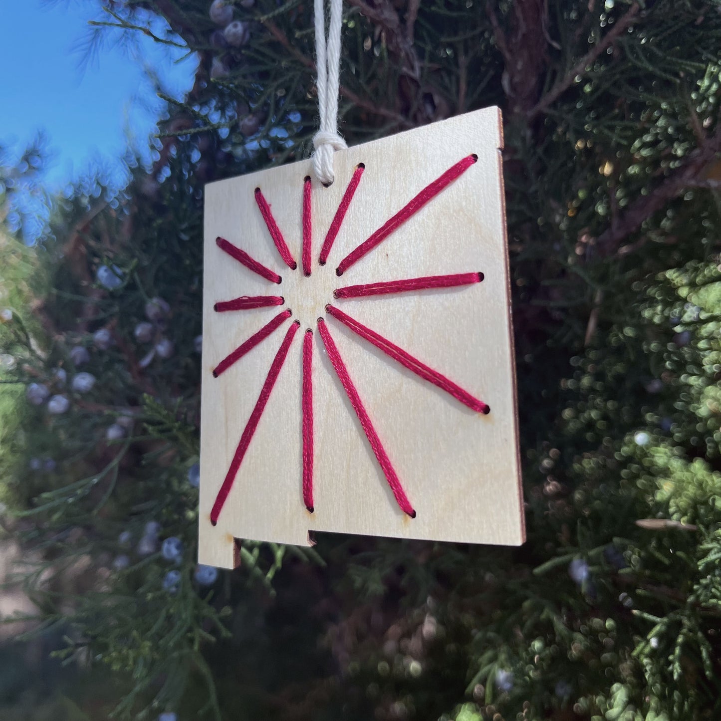 a laser cut wood ornament in the shape of New Mexico, hand embroidered in red thread with sunbeams coming out of a negative space heart, with an off white cord loop hanging on a pine tree