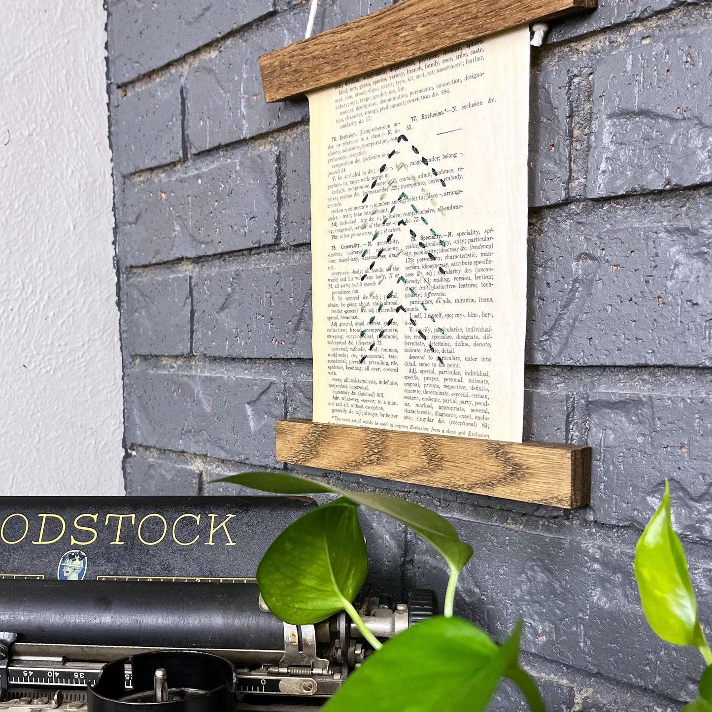 a paper wall hanging in a magnetic wood frame, made from a vintage thesaurus page, embroidered with the shape of a Christmas tree made from running stitches forming a column of chevrons, in shades of pine green thread, hanging on a grey brick wall with a typewriter and pothos plant in the foreground