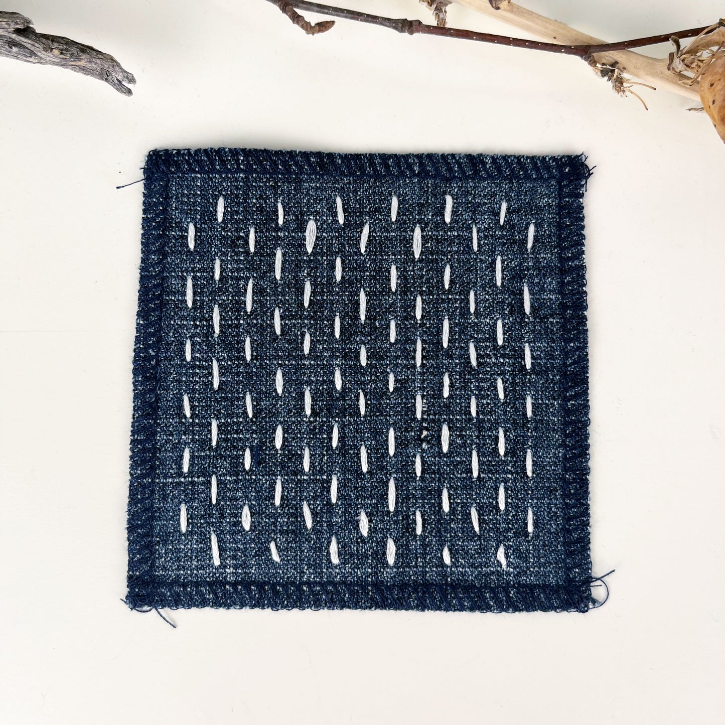 square denim patch with sashiko style running stitches in ivory on a white counter with dried twigs in the background
