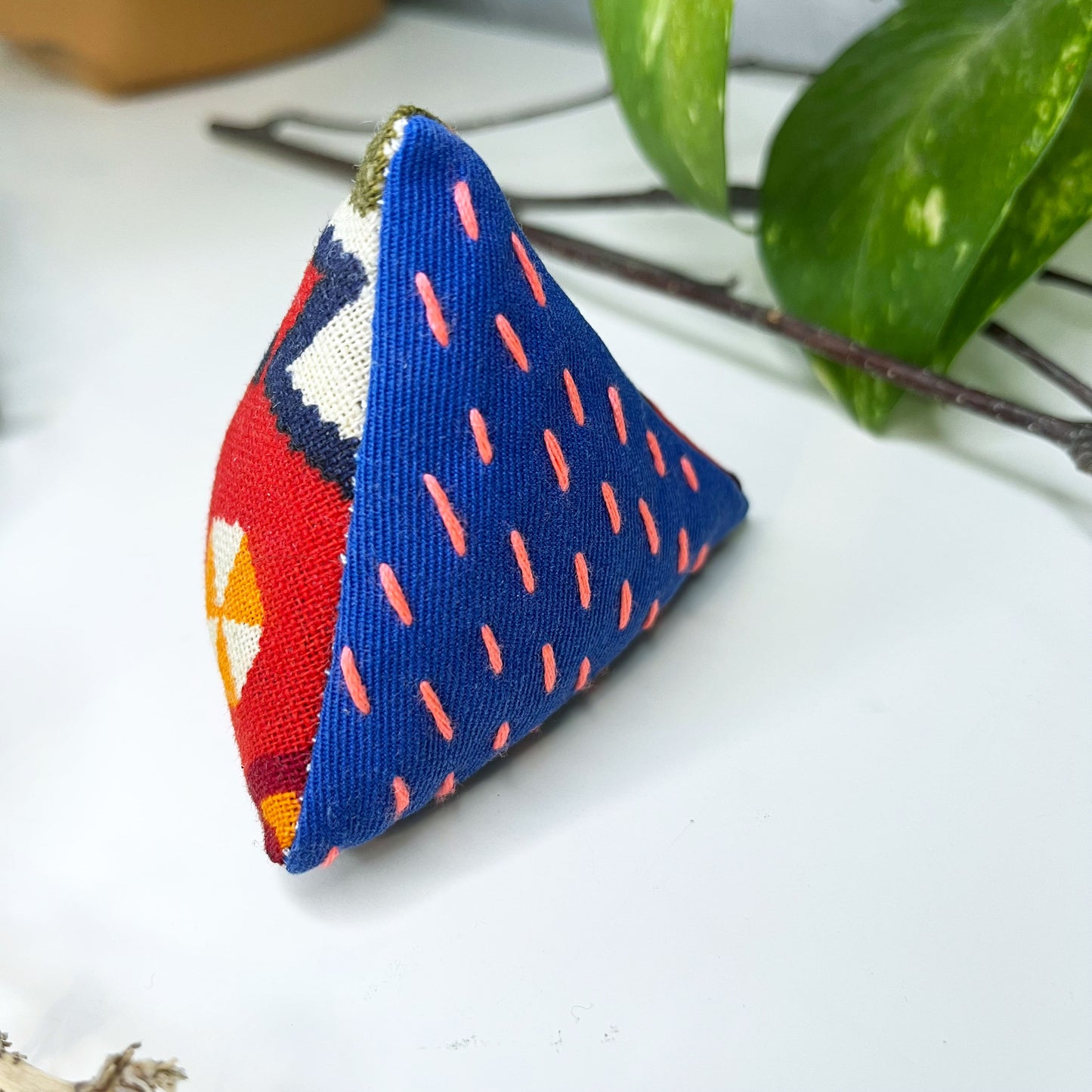 pyramid shaped pincushion made from fabrics in blue and a multi colored pattern, with sashiko running stitches on the blue in neon pink, on a dresser surrounded by plants