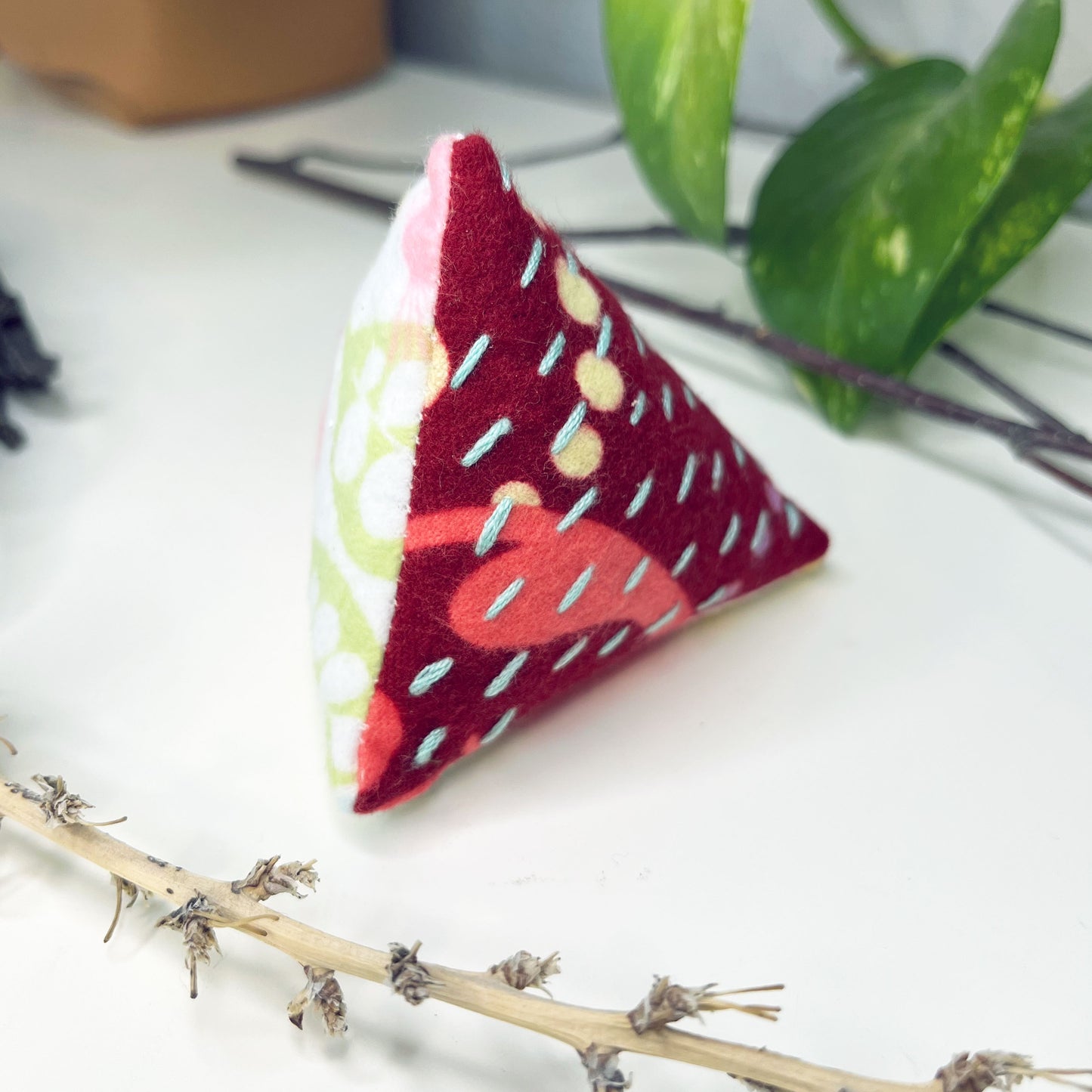 pyramid shaped pincushion made from patterned flannel fabrics in maroon fuchsia and white, with sashiko running stitches on them in blue, on a dresser surrounded by plants