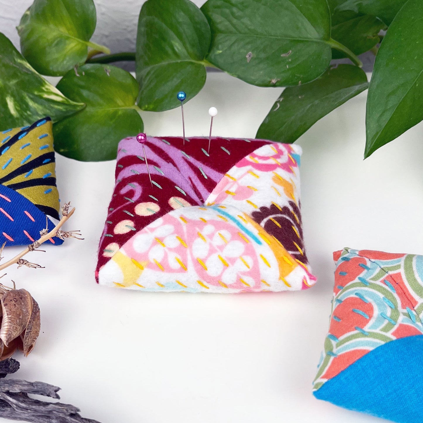 square shaped pincushion with pins in it, made from two colorful fabrics with diagonal seams, with sashiko stitching in contrasting colors, on a white background