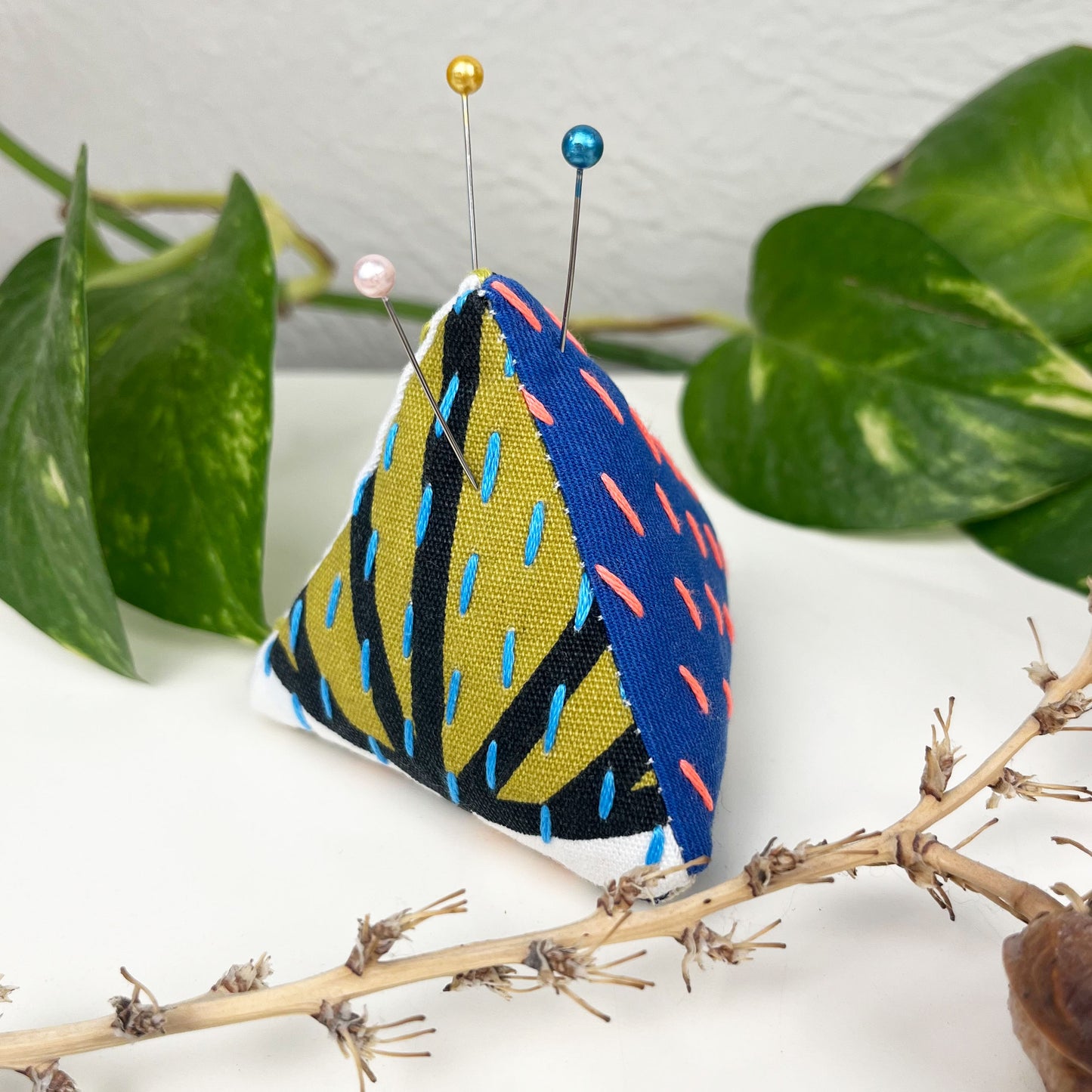 pyramid shaped pincushion made from patterned fabrics in blue chartreuse white and black, with sashiko running stitches on them in blue and coral, on a dresser surrounded by plants