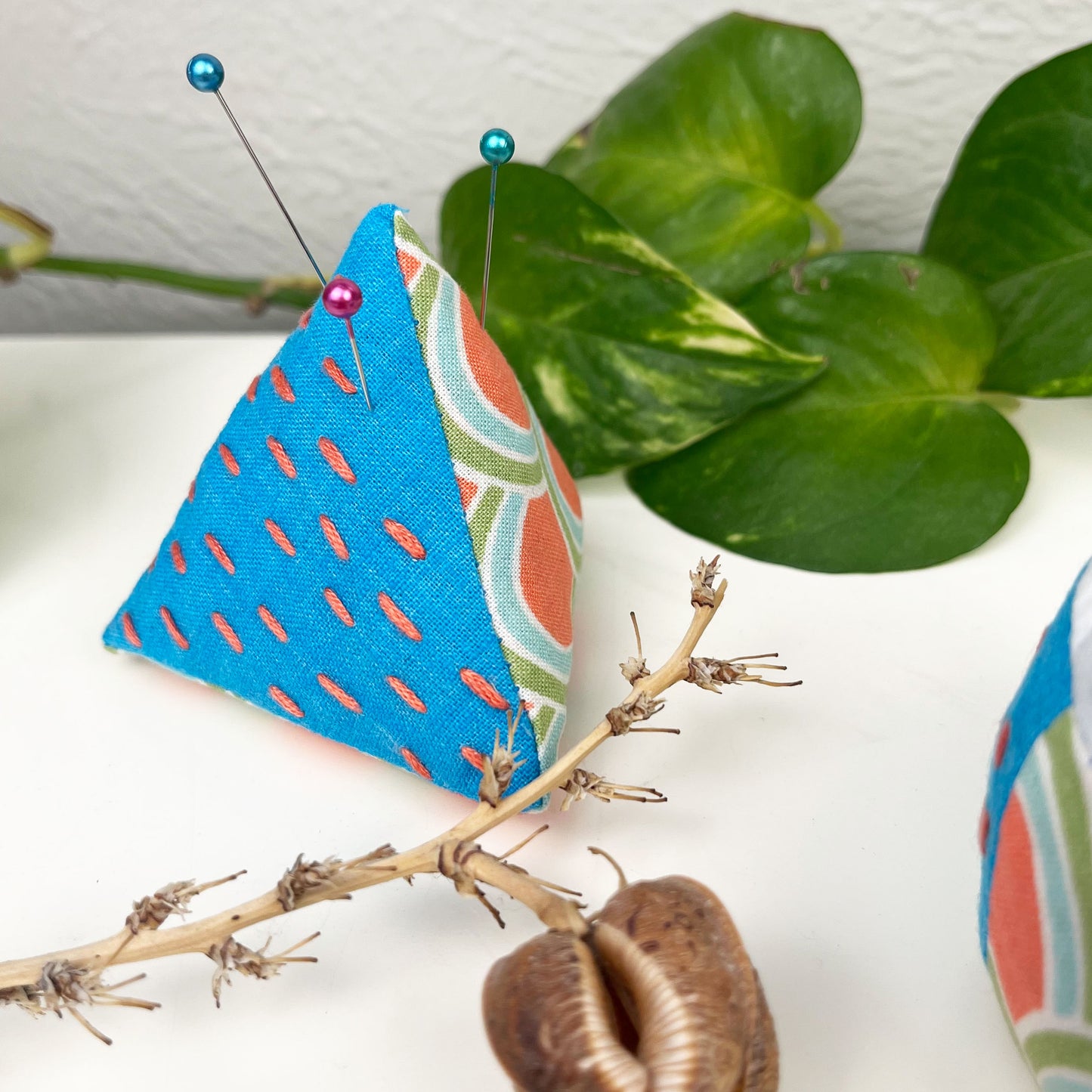 pyramid shaped pincushion made from patterned and solid fabrics in blue coral and green, with sashiko running stitches in coral, on a dresser surrounded by plants