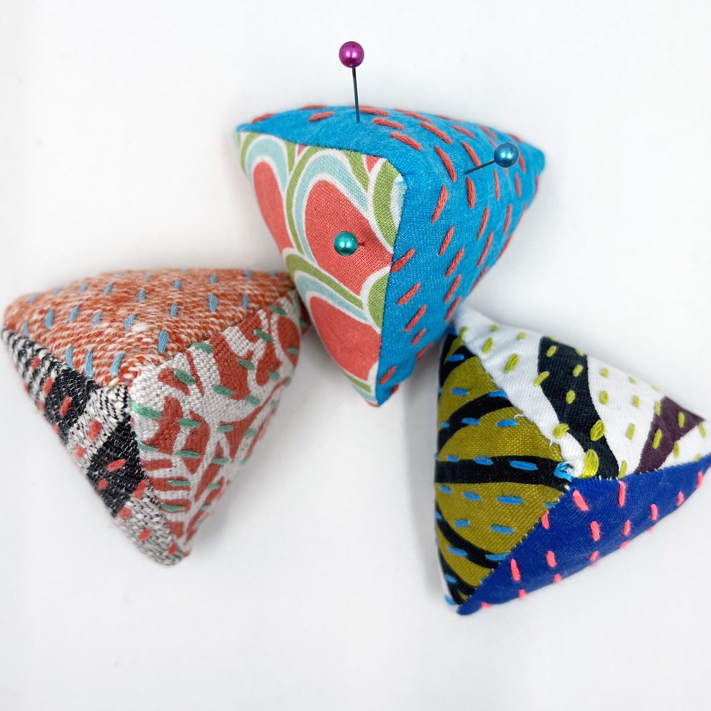 birds eye view of three pyramid shaped pincushions, made from different combinations of colorful fabric, some with sashiko stitching on contrasting colors, on a white background