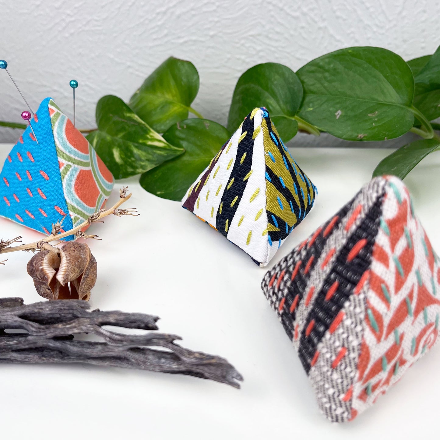 three pyramid shaped pincushions, made from different combinations of colorful fabric, some with sashiko stitching on contrasting colors, on a white dresser with a pothos plant and dried plants around it