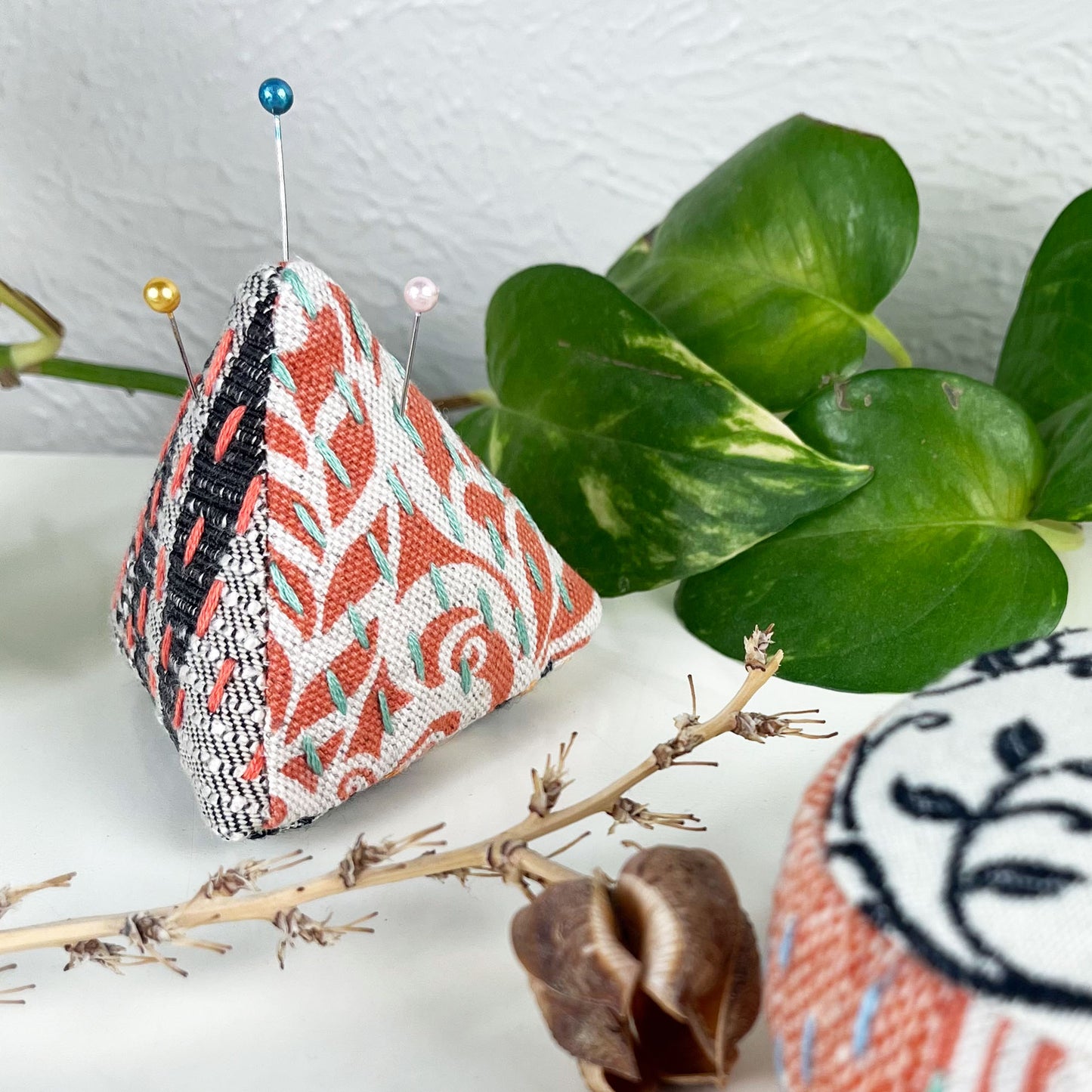 pyramid shaped pincushion made from patterned fabrics in black grey orange and cream, with sashiko running stitches on them in orange and green, on a dresser surrounded by plants