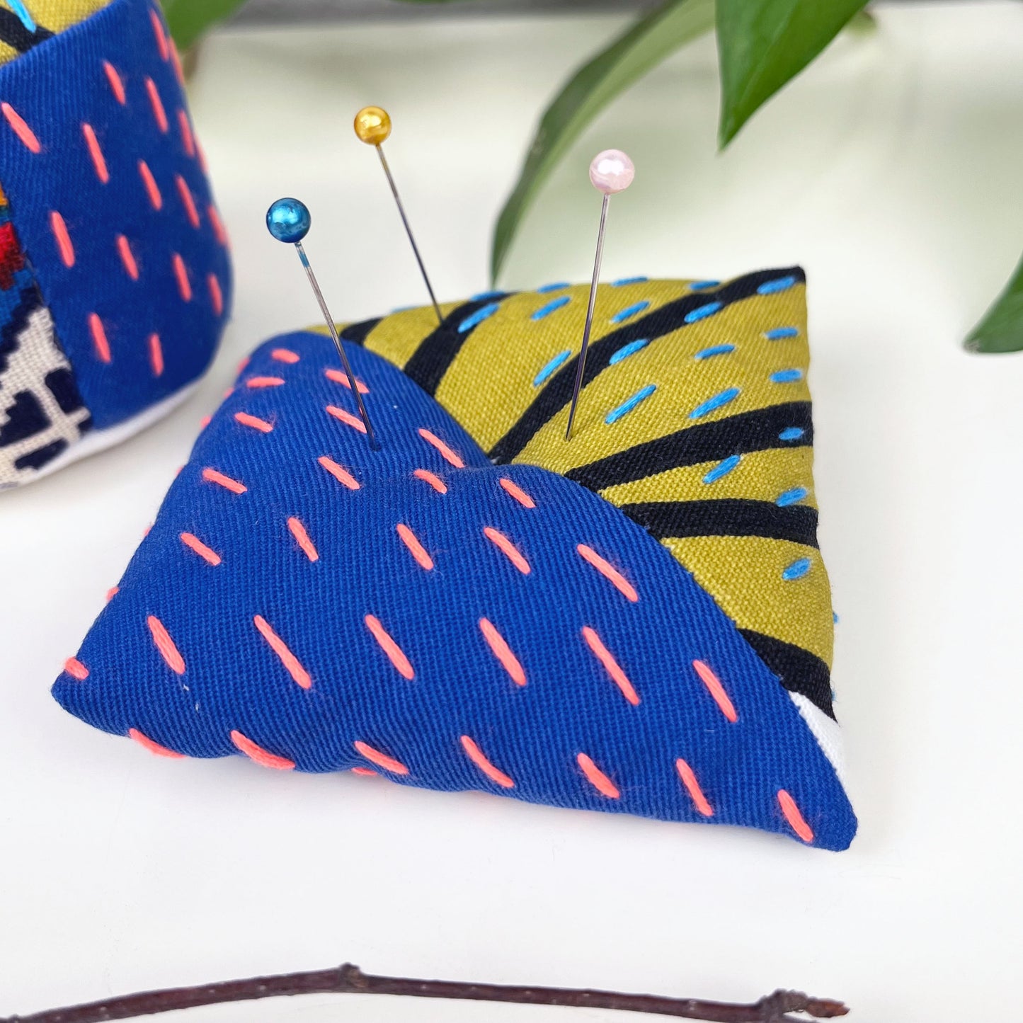 square shaped pincushion with pins in it, made from two colorful fabrics with diagonal seams, with sashiko stitching in contrasting colors, on a white background