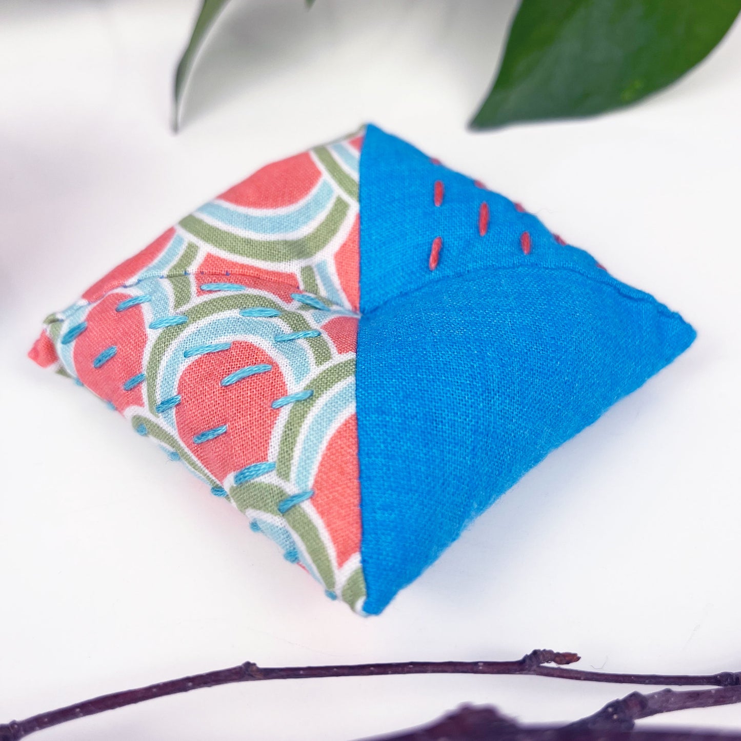 square shaped pincushion with pins in it, made from two colorful fabrics with diagonal seams, with sashiko stitching in contrasting colors, on a white background