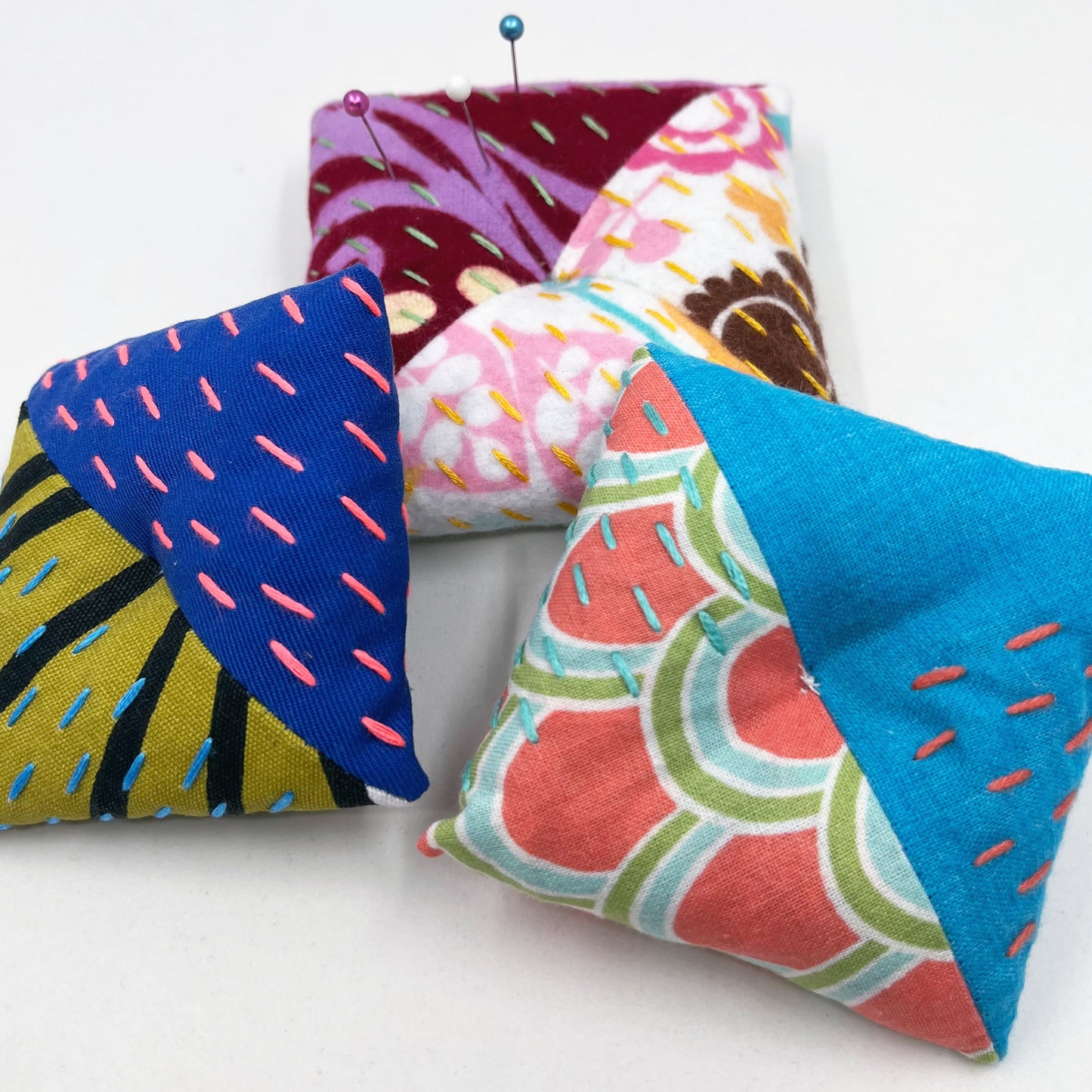three square shaped pincushions, made from different combinations of colorful fabric with diagonal seams, with sashiko stitching in contrasting colors, on a white background