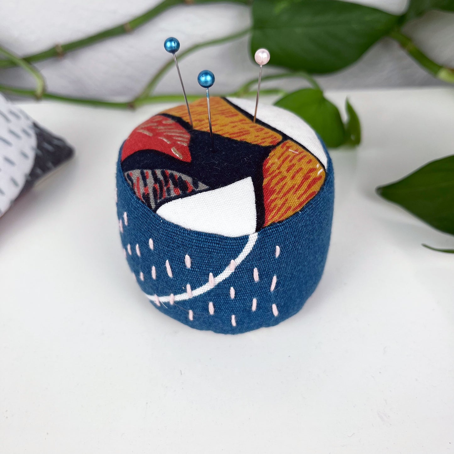 a round pincushion made from multi colored floral and teal fabric with contrasting sashiko stitches on the sides