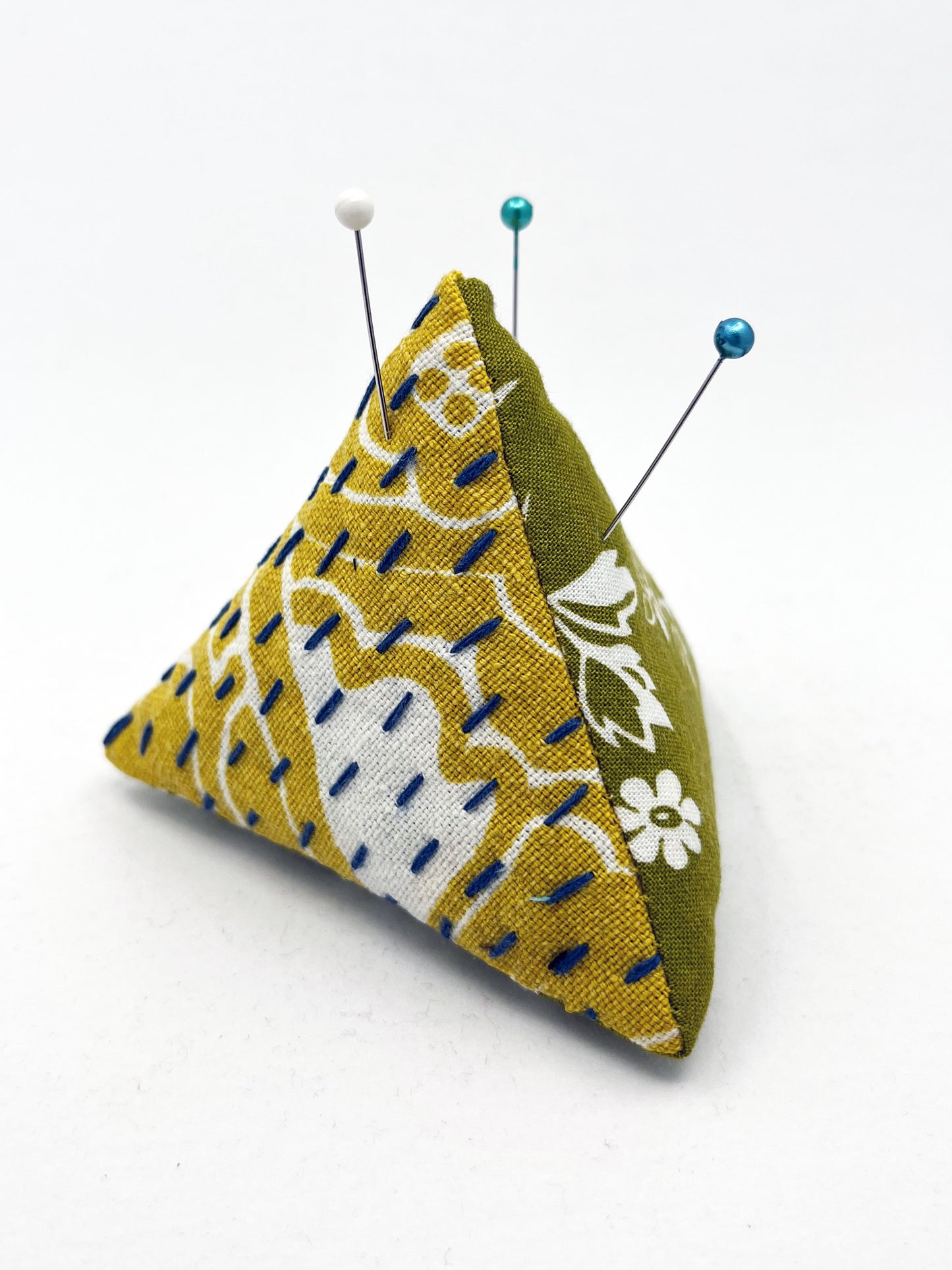 a pyramid pincushion made from floral olive and mustard colored fabric with contrasting sashiko stitches on a white counter
