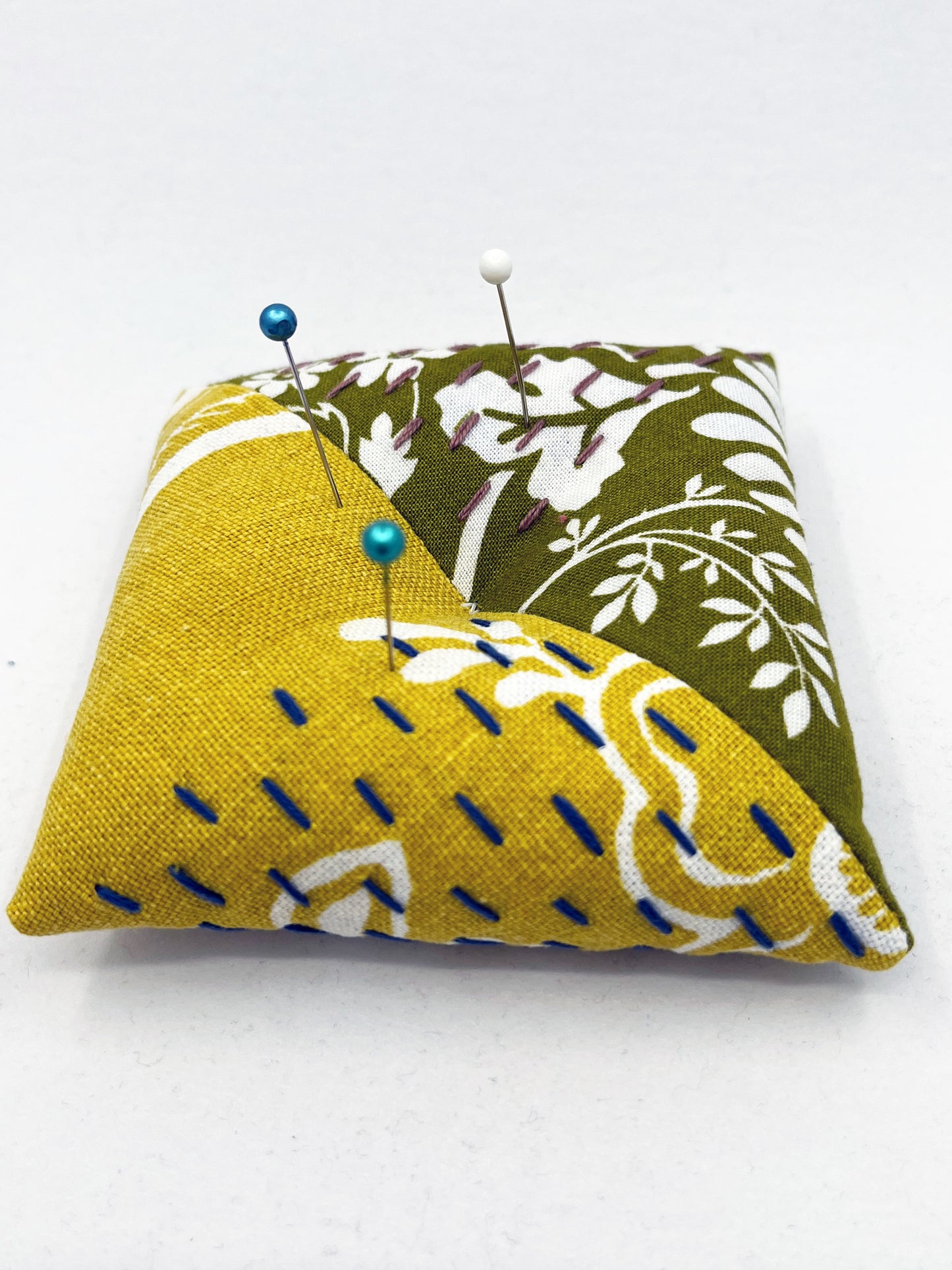 a square pillow shaped pincushion made from floral olive and mustard colored fabric with contrasting sashiko stitches on a white counter