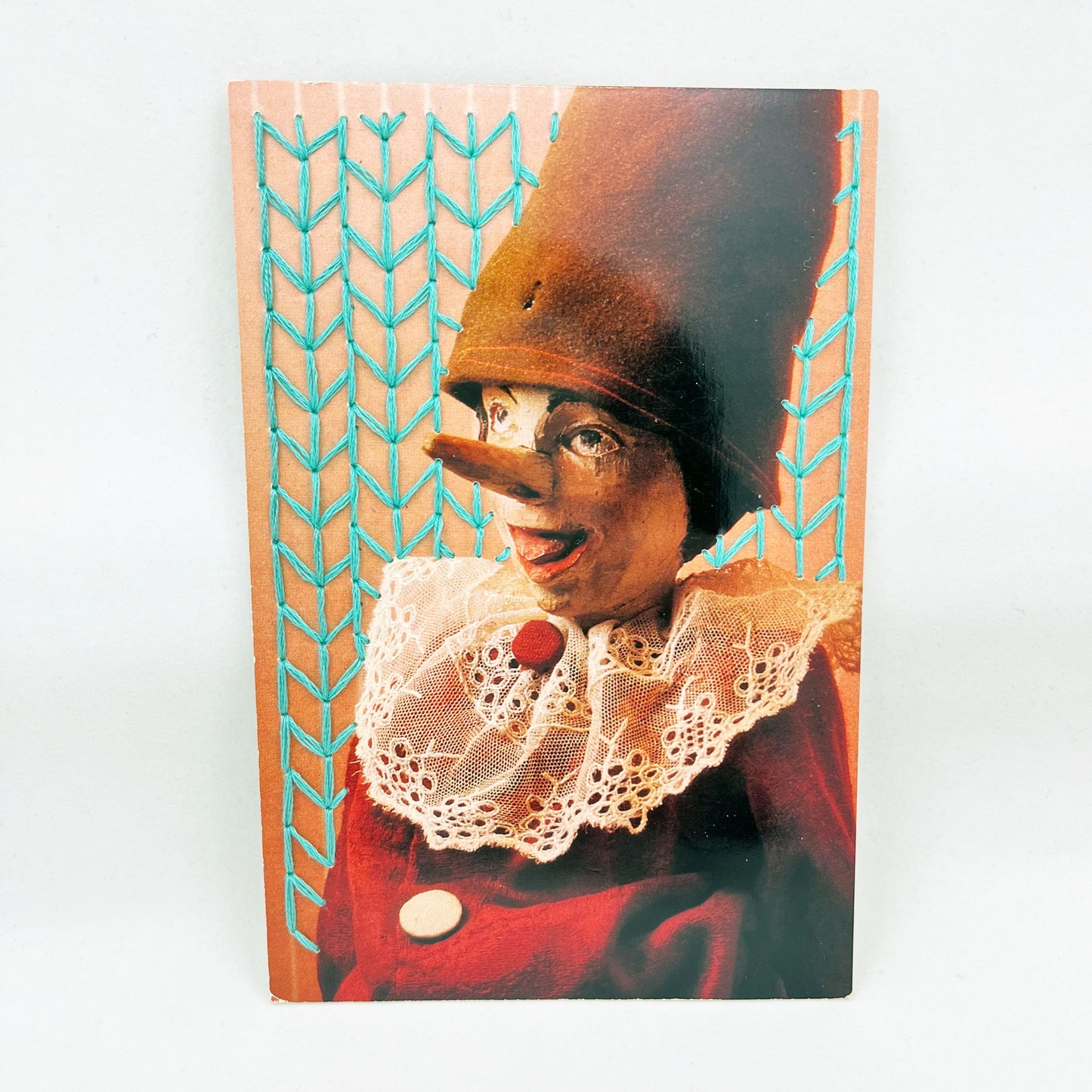 postcard of an old Pinocchio puppet, with the background hand embroidered in aqua in an all over chevron diamond pattern, on a white background