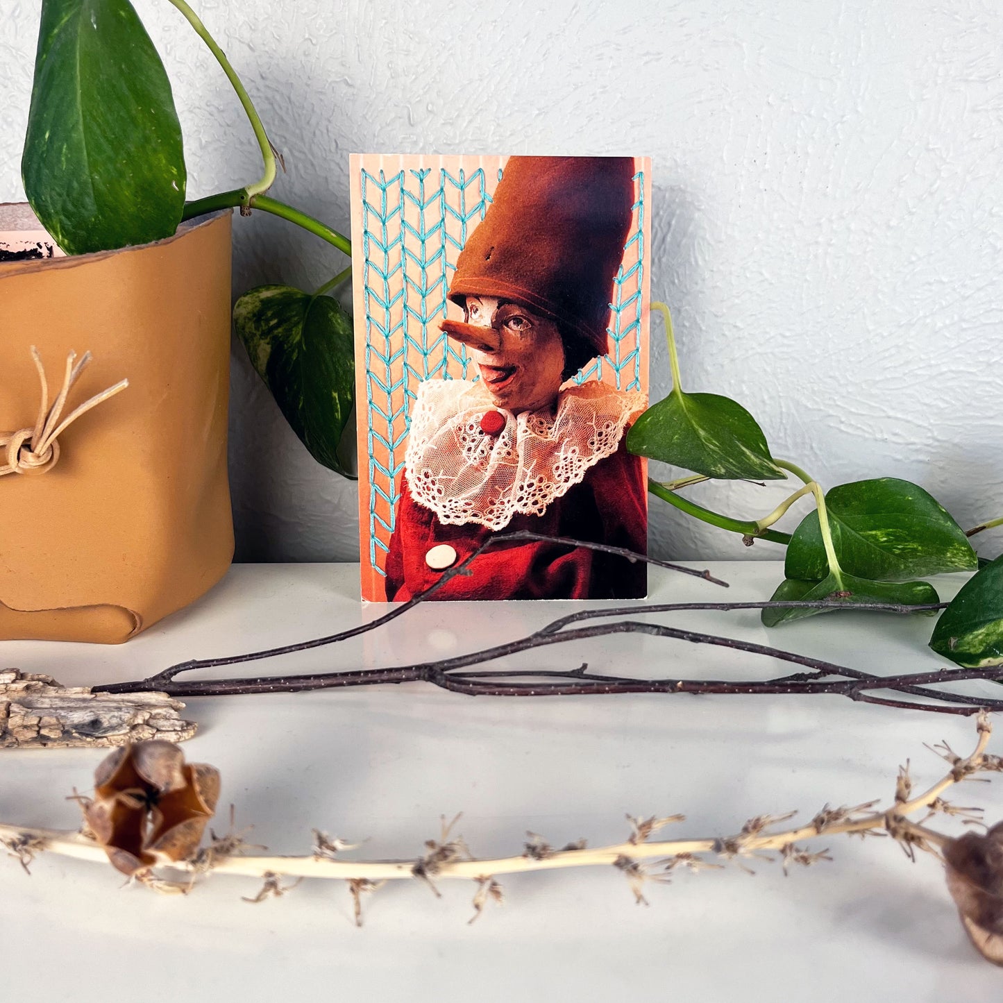 postcard of an old Pinocchio puppet, with the background hand embroidered in aqua in an all over chevron diamond pattern, the postcard has a plant vine behind it, and twigs on the counter in front of it