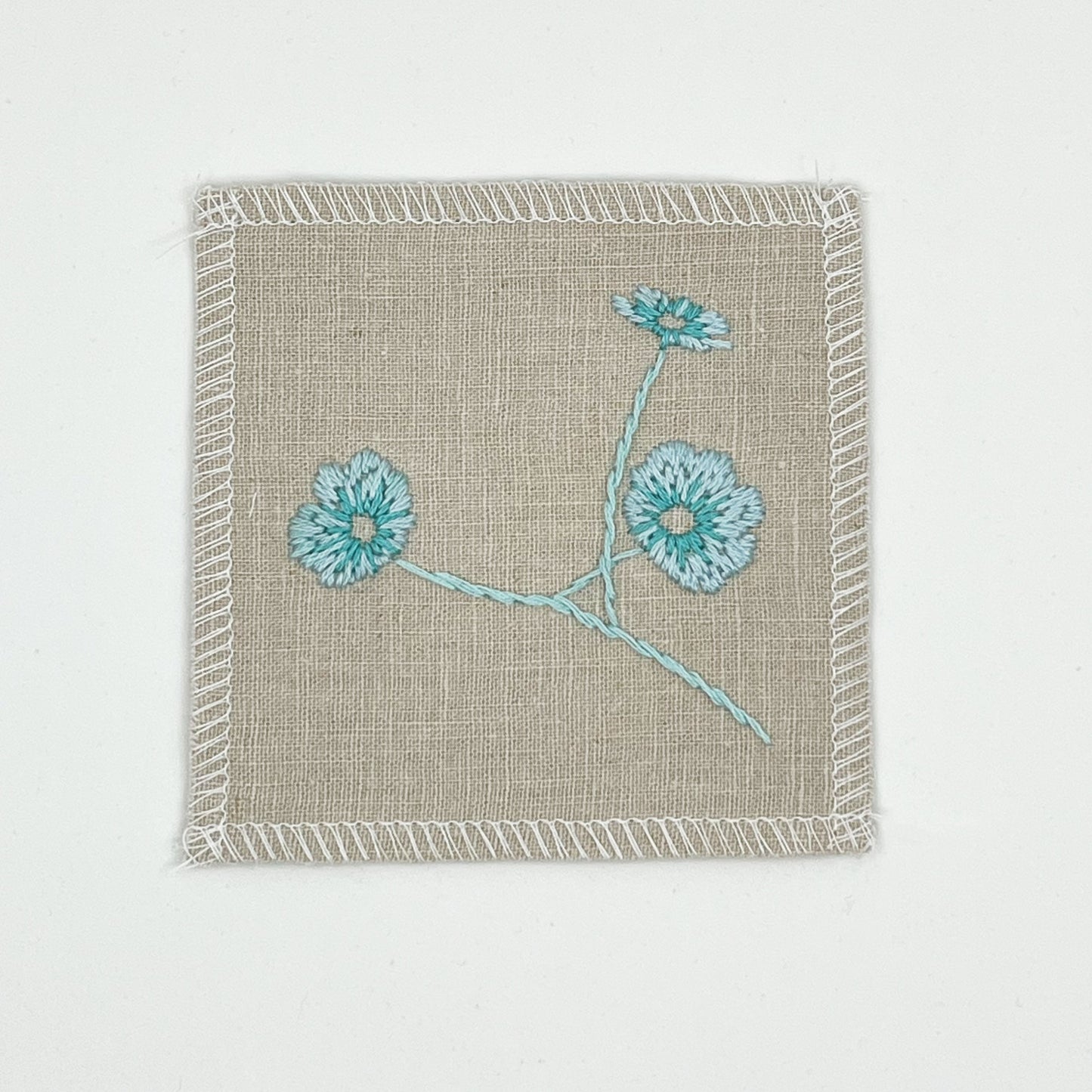 a natural colored square patch, hand embroidered with three poppies on a stem, in shades of robin's egg blue, with overlocked edges, on a white background