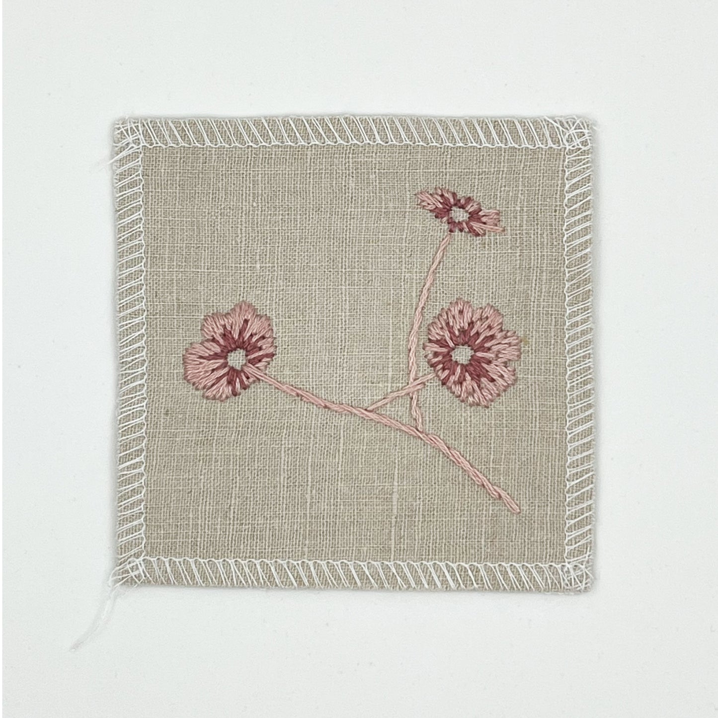 a natural colored square patch, hand embroidered with three poppies on a stem, in shades of pink, with overlocked edges, on a white background