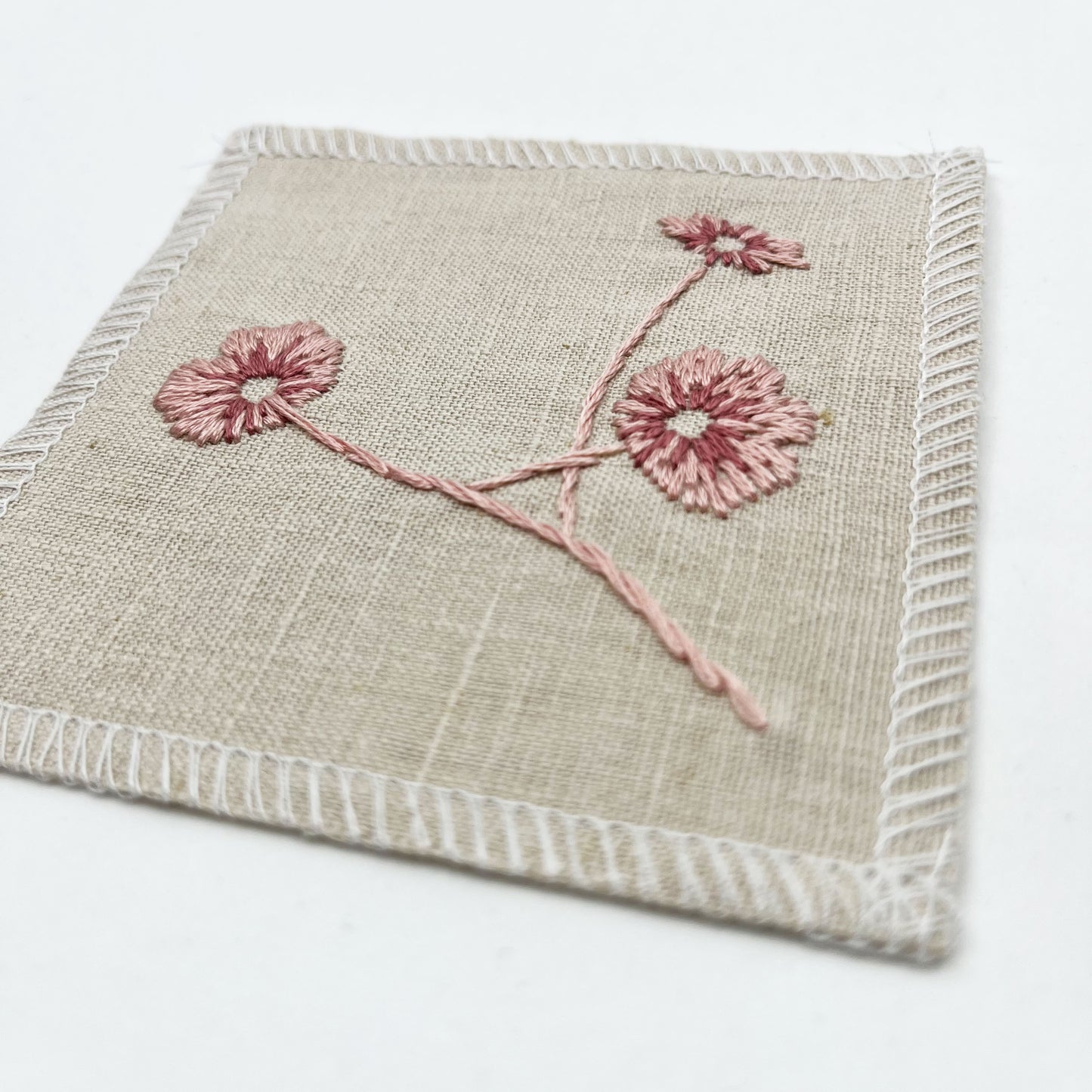 a close up angled view of a natural colored square patch, hand embroidered with three poppies on a stem, in shades of pink, with overlocked edges, on a white background