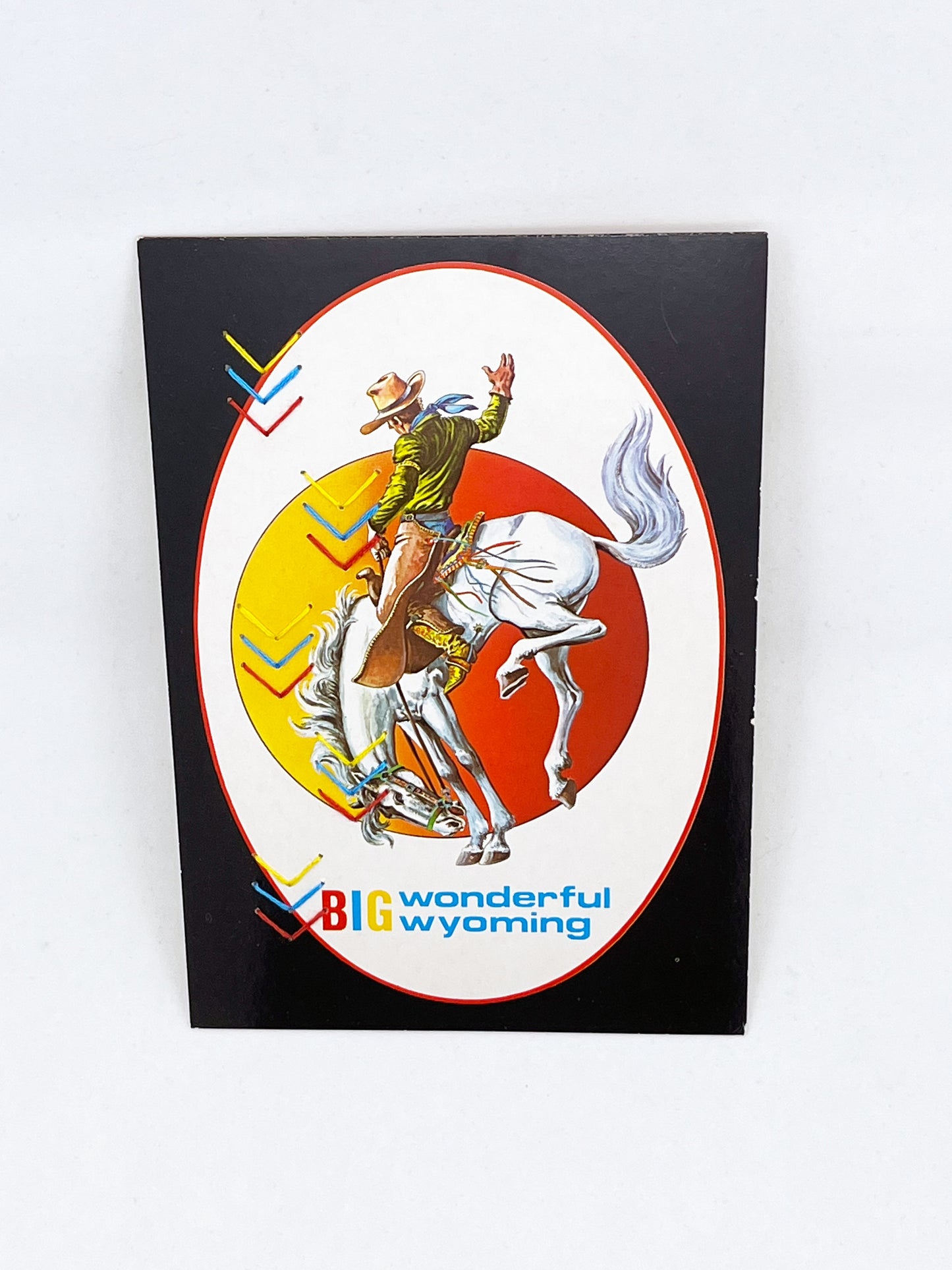 a vintage postcard of a cowboy on a bucking horse with the words big wonderful Wyoming below them hand embroidered over with groups of yellow blue and red chevrons in two alternating rows down the left side