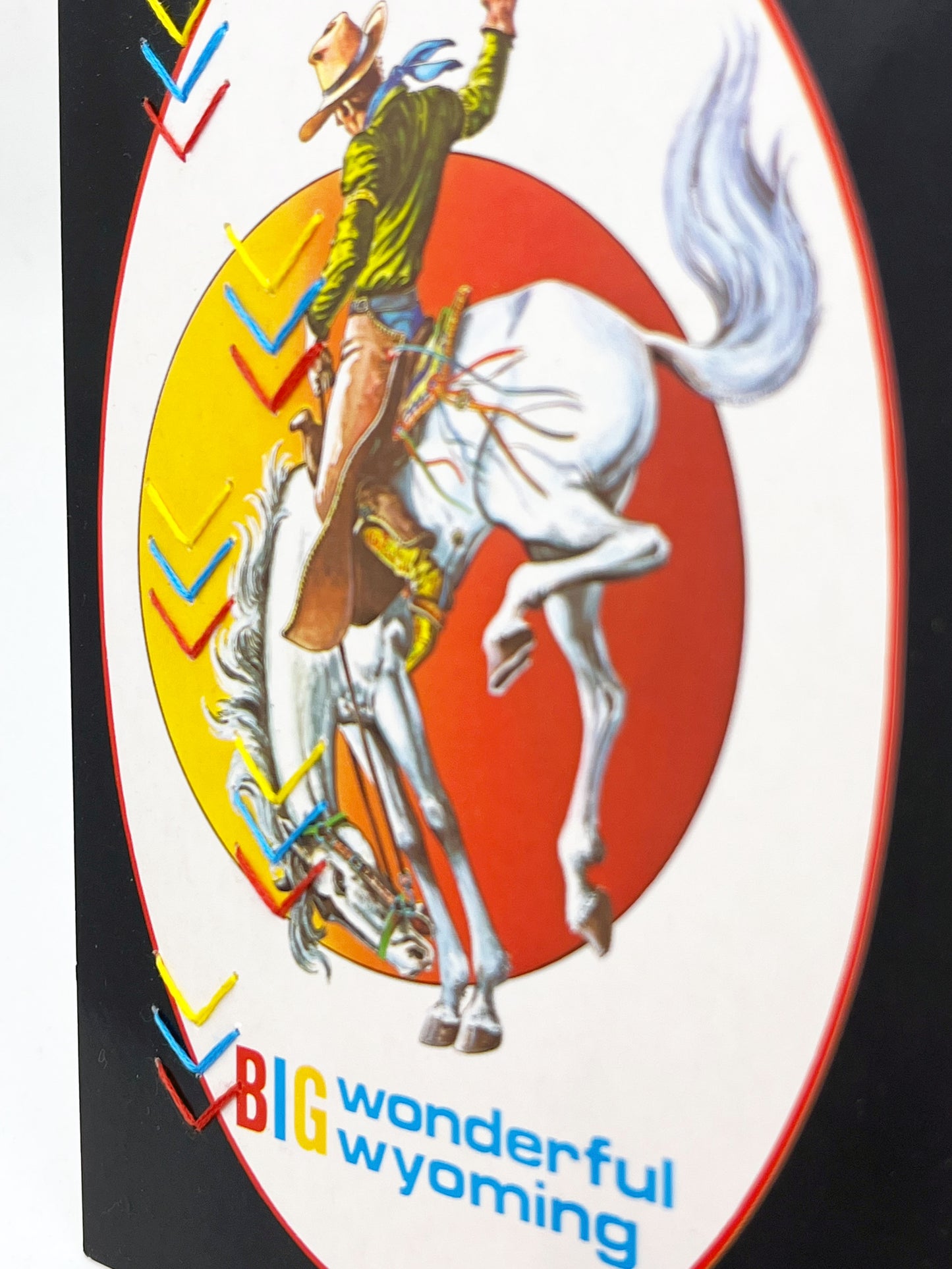 a vintage postcard of a cowboy on a bucking horse with the words big wonderful Wyoming below them hand embroidered over with groups of yellow blue and red chevrons in two alternating rows down the left side