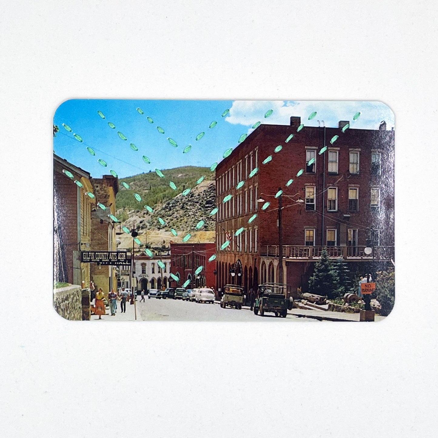 a vintage postcard of a small mountain town in Colorado, hand stitched with four rows of chevrons in blue green floss, on a white background