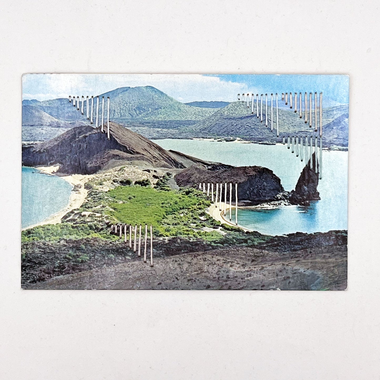 an old postcard of part of Galapagos Ecuador, with hand embroidered triangles randomly placed in pastel greens blues peach and ivory