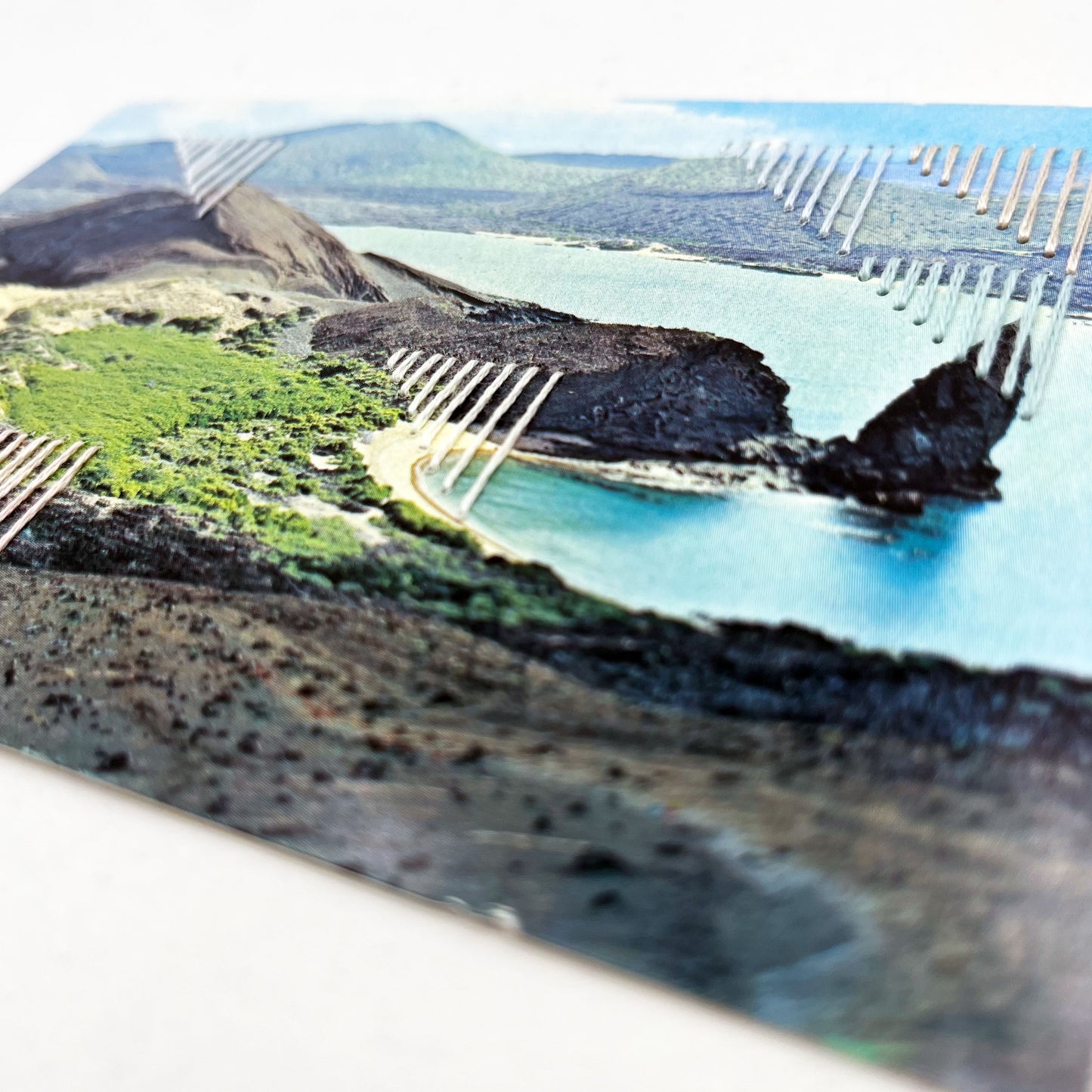 close up angled view an old postcard of part of Galapagos Ecuador, with hand embroidered triangles randomly placed in pastel greens blues peach and ivory