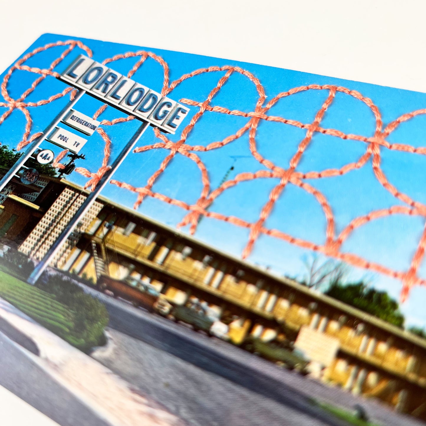 close up angled view of an old postcard of a motel called Lorlodge, with a hand embroidered circle and line design like breeze blocks in peach stitched in the background on the sky only, the postcard is sitting on a white background