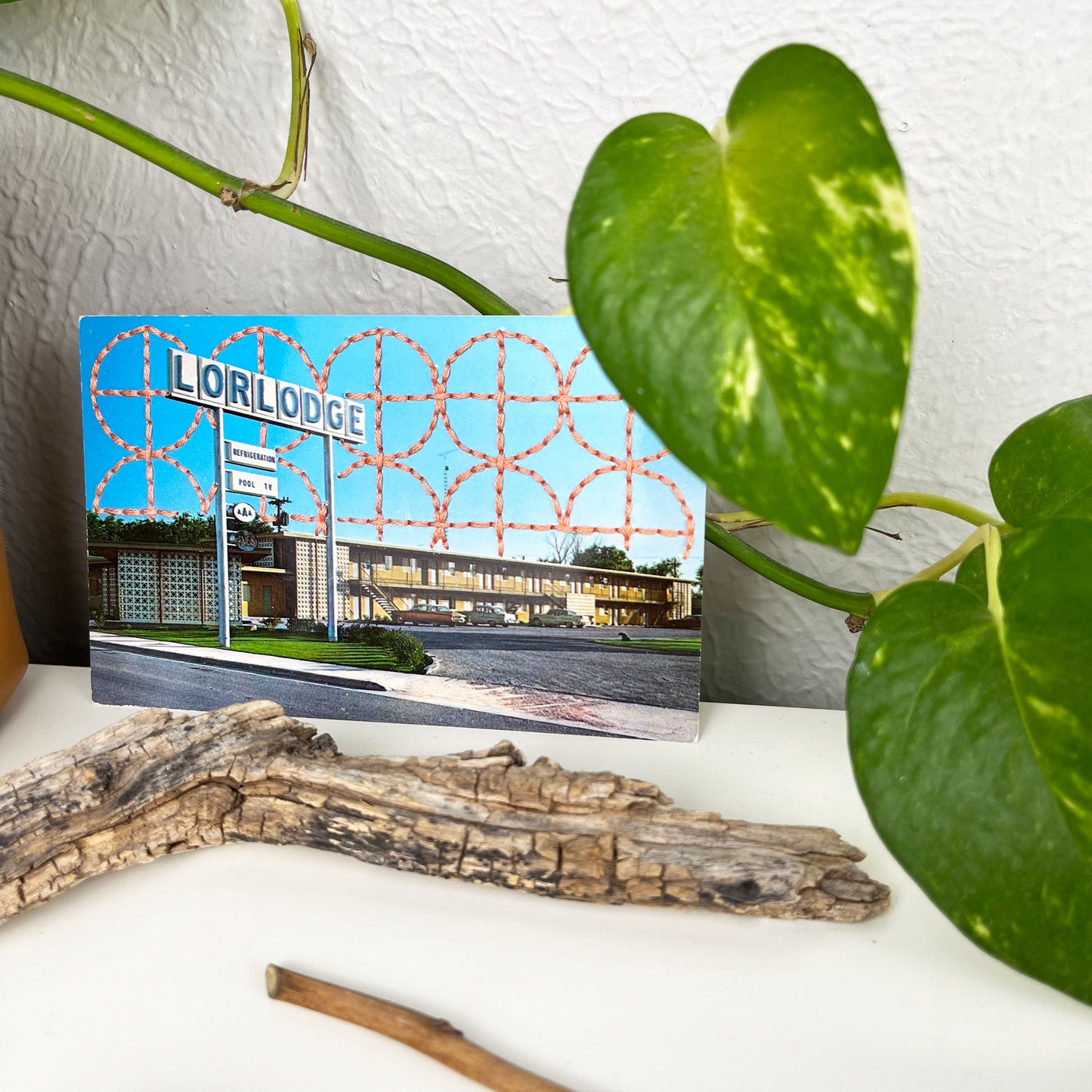 an old postcard of a motel called Lorlodge, with a hand embroidered circle and line design like breeze blocks in peach stitched in the background on the sky only, the postcard is sitting on a white dresser with a plant vine behind it and sticks and dried flowers in front of it