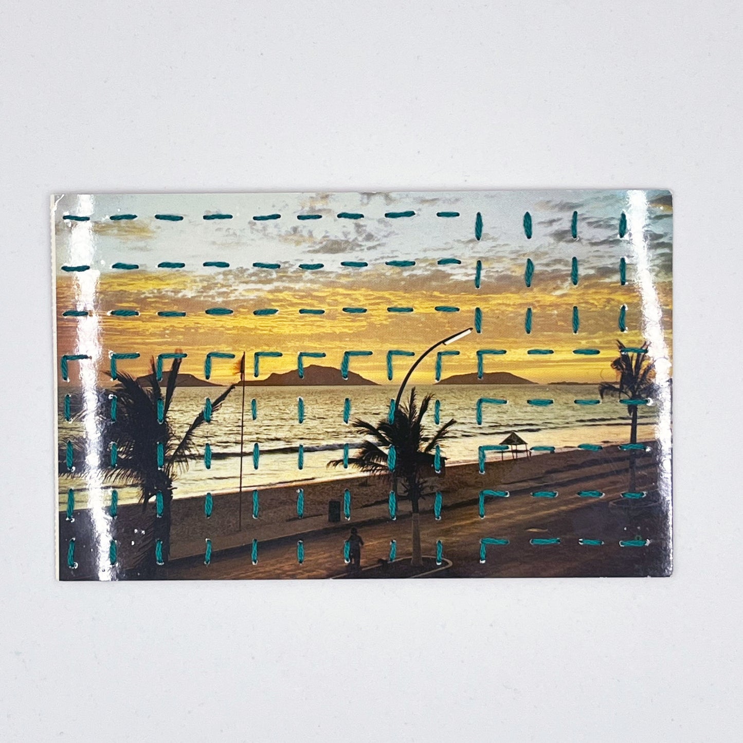 a vintage postcard of a beach sunset in Mazatlan Mexico, hand stitched over with a basketweave running stitch pattern in teal