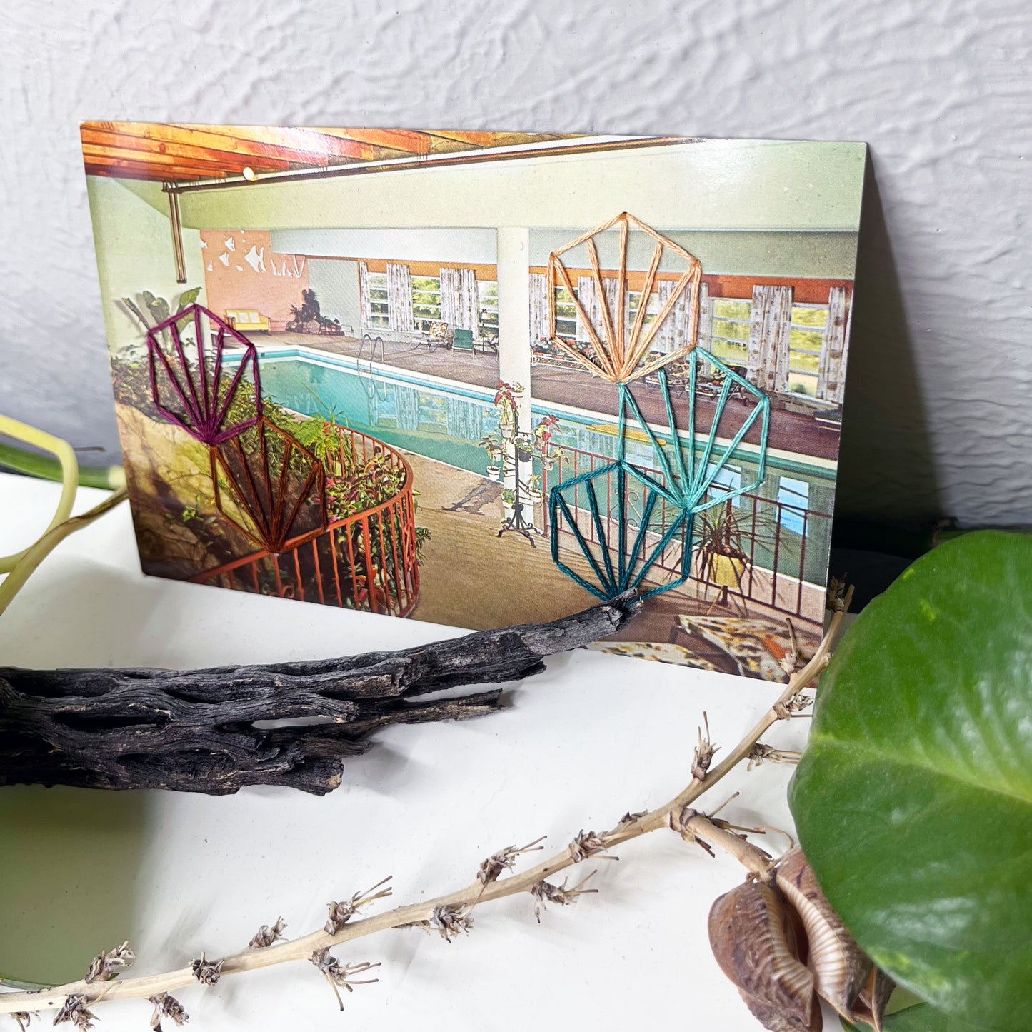 vintage postcard of an indoor pool, with art deco hexagons hand embroidered on it in aqua teal plum peach and brown, sitting on a white surface with a pothos plant and dried cactus and yucca seed pods around it