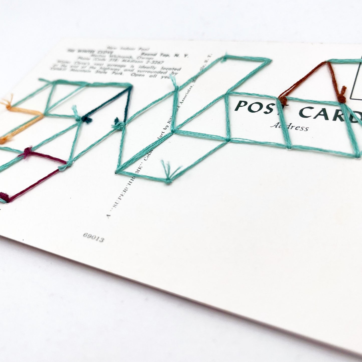 back side of a vintage postcard of an indoor pool, with a grid of triangles hand embroidered on it in aqua teal plum peach and brown