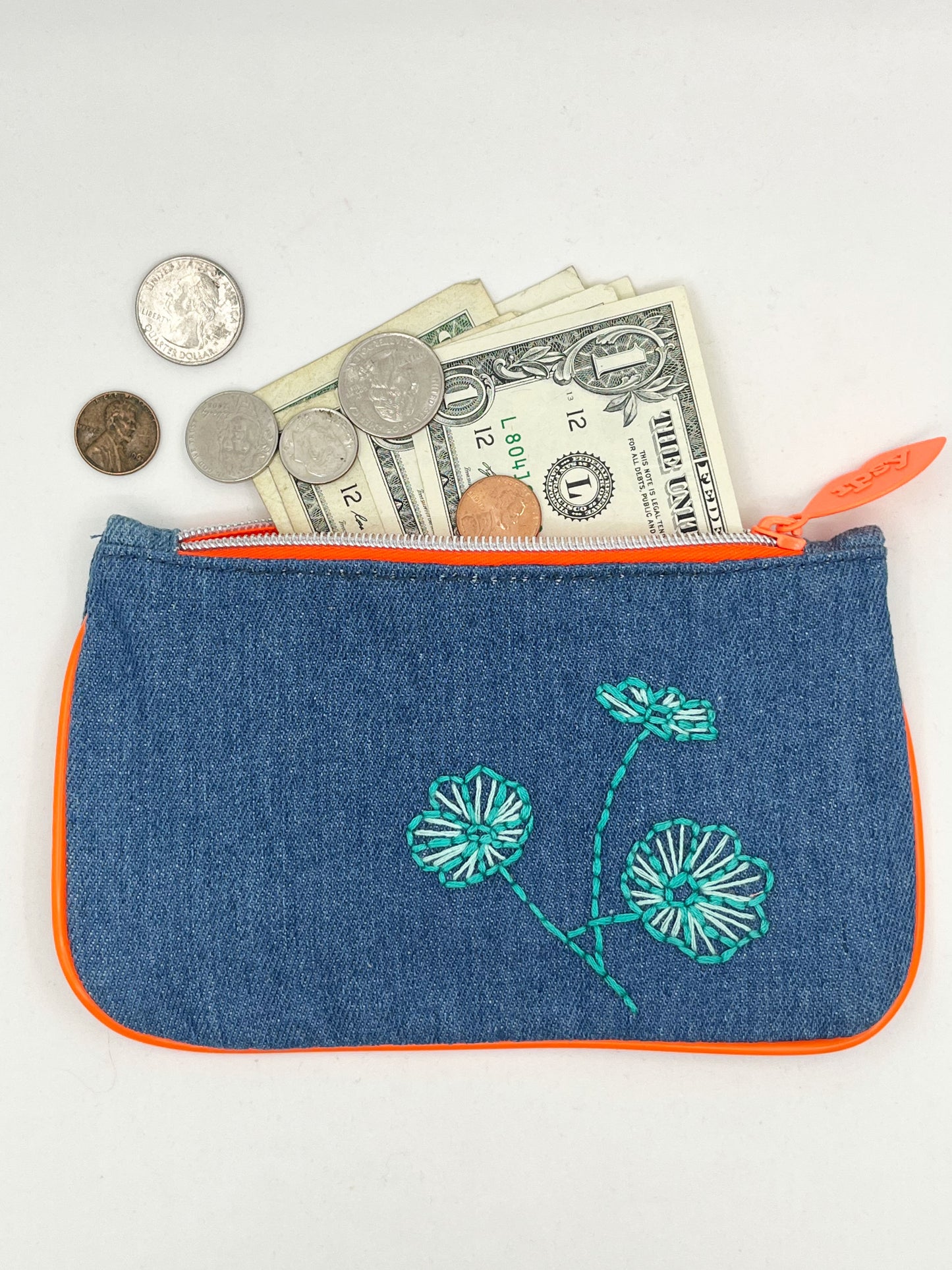denim Ipsy zip pouch with neon orange piping and zipper, hand embroidered with aqua poppies with money coming out of the top of it
