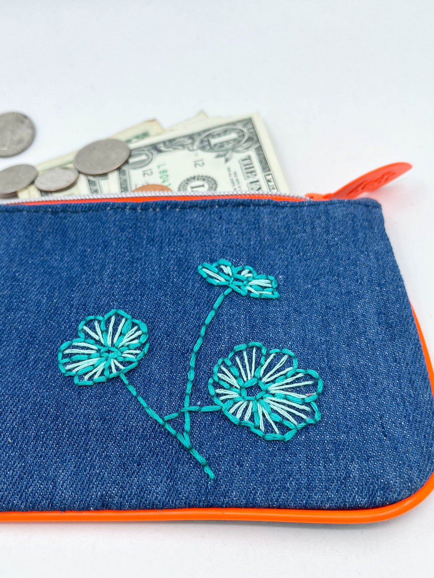 denim Ipsy zip pouch with neon orange piping and zipper, hand embroidered with aqua poppies with money coming out of the top of it