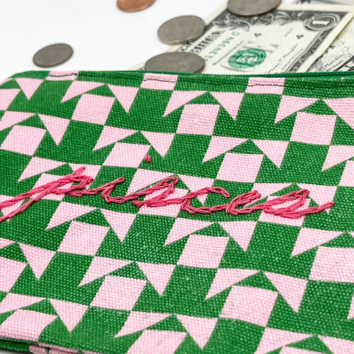 zip pouch with a pink and green geometric design on it hand embroidered in hot pink with the word pisces in chainstitch with money spilling out the top of it