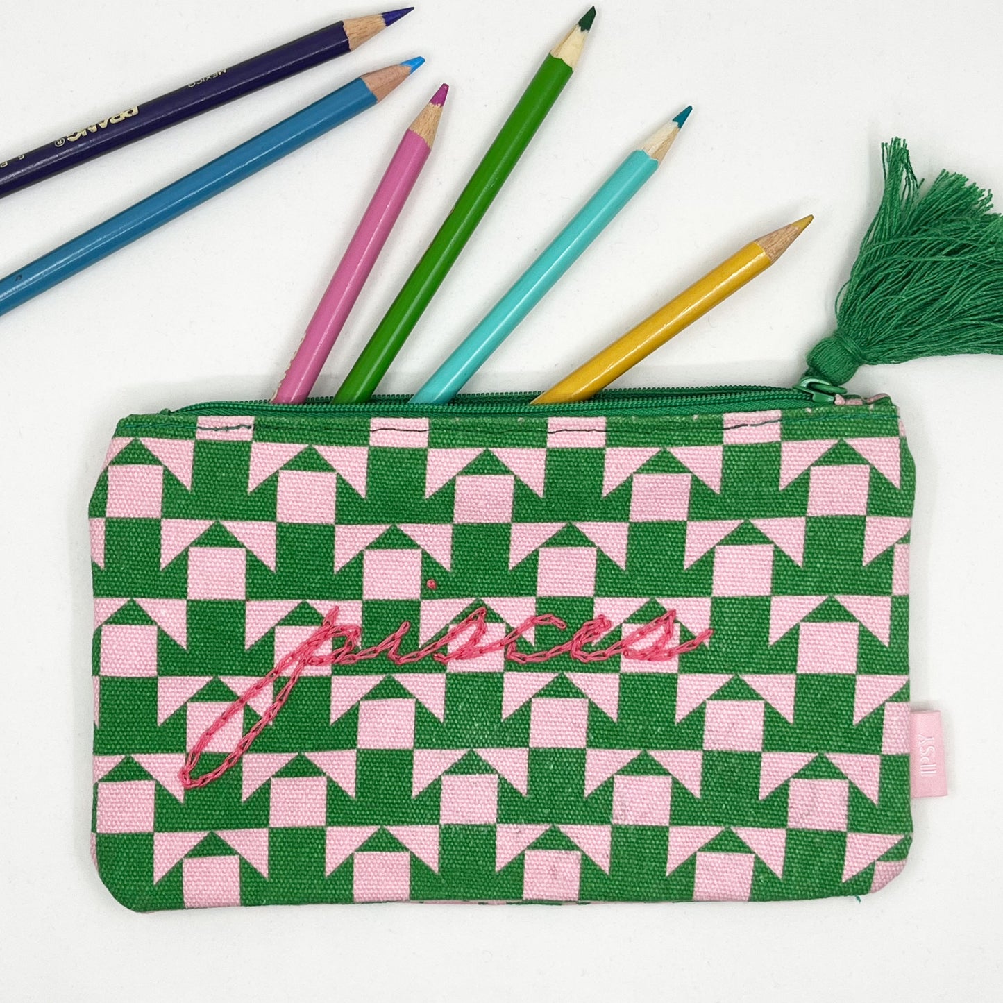 zip pouch with a pink and green geometric design on it hand embroidered in hot pink with the word pisces in chainstitch with colored pencils spilling out the top of it
