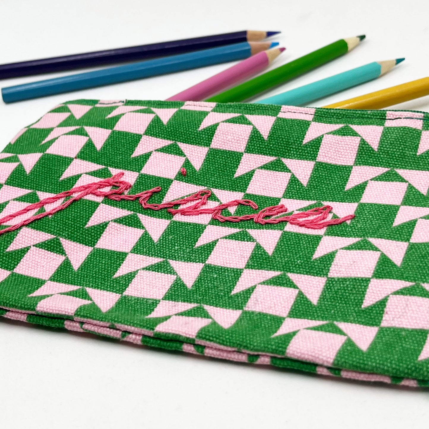 zip pouch with a pink and green geometric design on it hand embroidered in hot pink with the word pisces in chainstitch with colored pencils spilling out the top of it