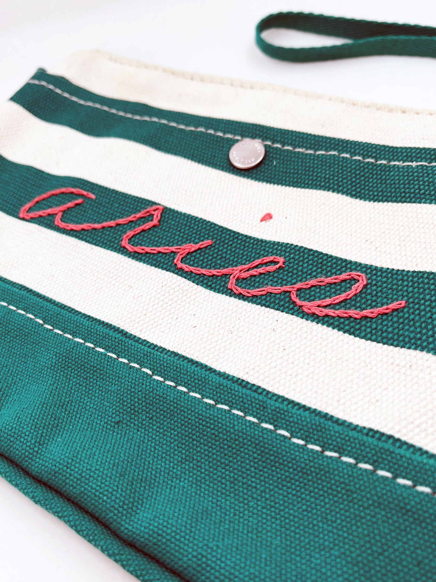 close up angled view of green and cream zipper wristlet with the word aries hand embroidered on it in coral chainstitch