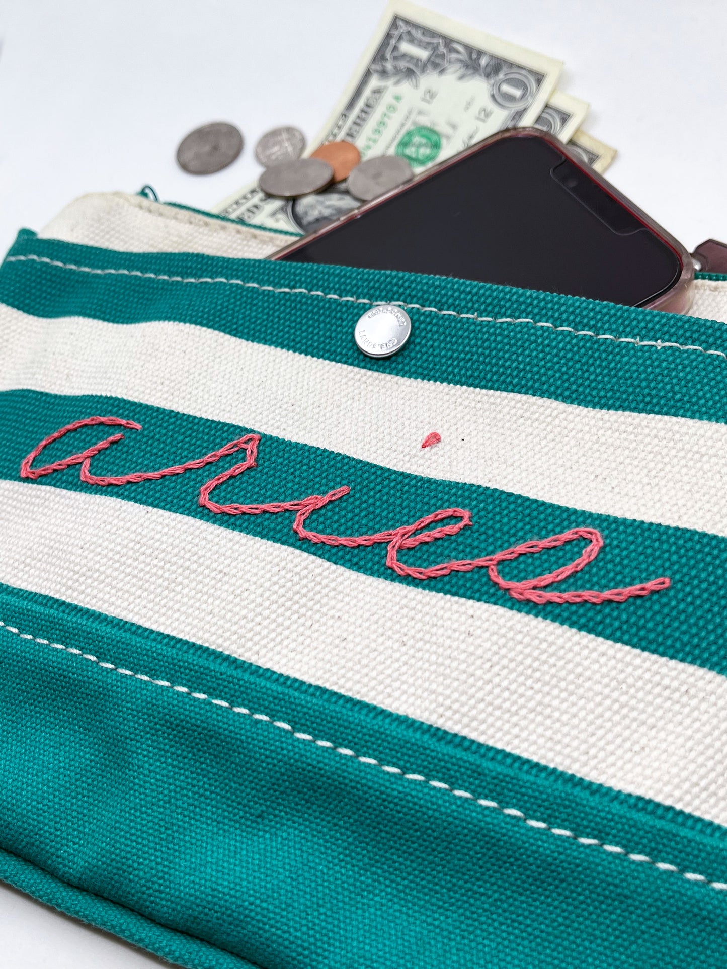 close up angled view of green and cream zipper wristlet with the word aries hand embroidered on it in coral chainstitch with money and a phone coming out of the top of it