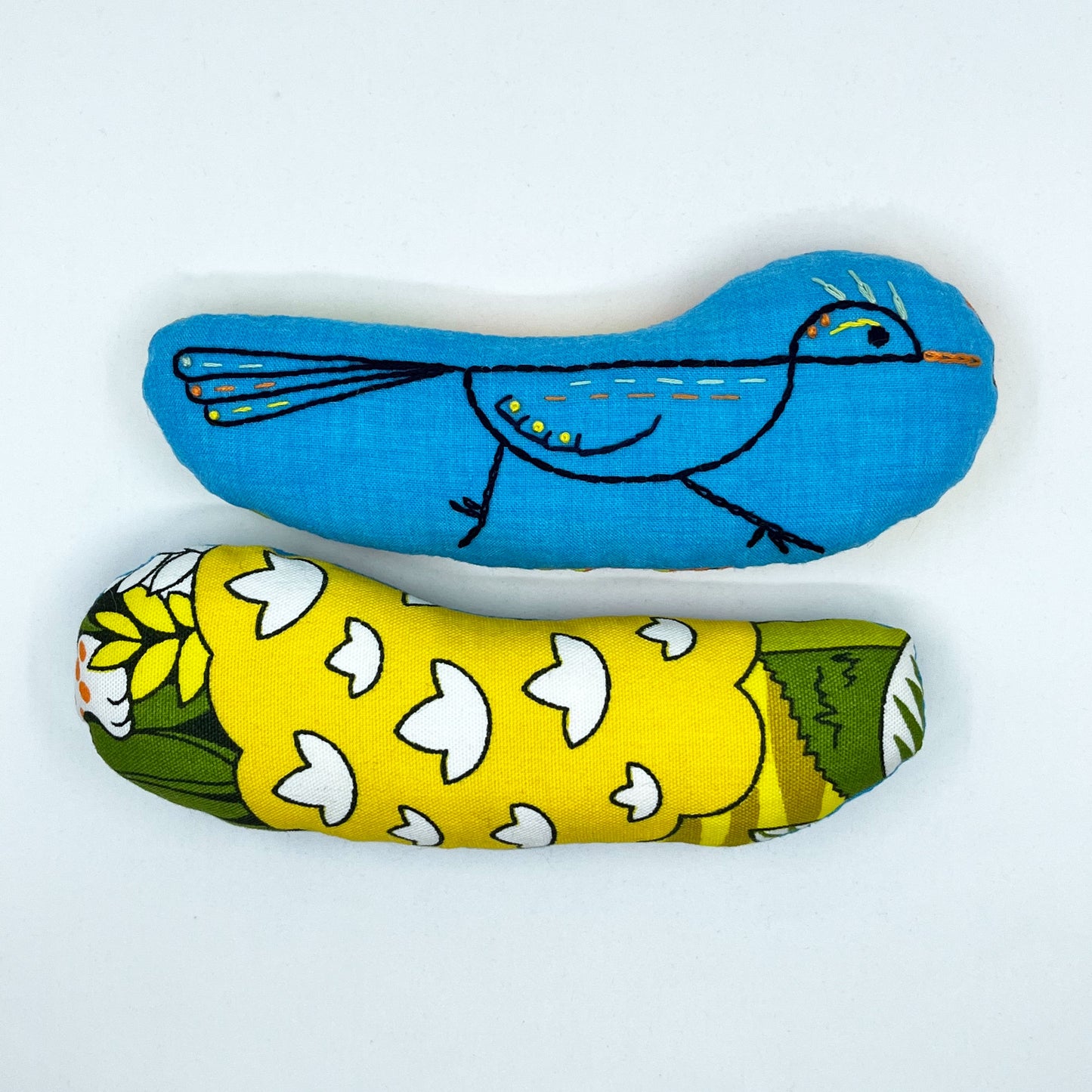 a colorfully hand embroidered stuffed pillow animal of a roadrunner on blue fabric, behind it one turned around showing the back side made of fabric with a cartoon jungle print