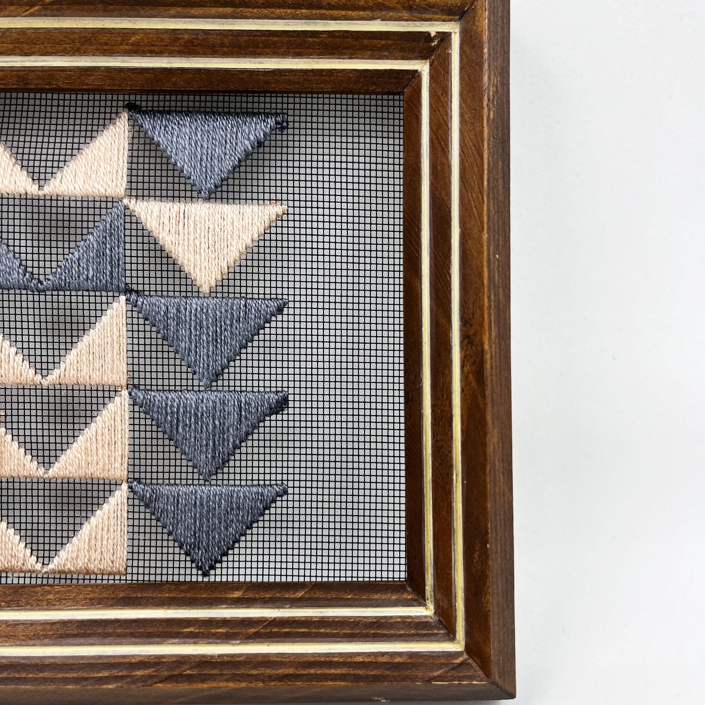 close up abstract half view of a piece of window screen hand stitched with 3 rows of triangles resembling the flying geese quilt blocks, in grey and peach, in a wood frame, on a white background