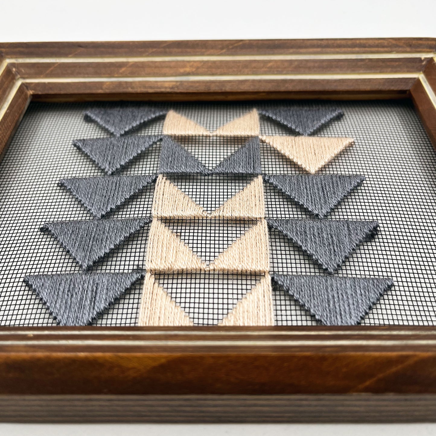 close up angled view of a piece of window screen hand stitched with 3 rows of triangles resembling the flying geese quilt blocks, in grey and peach, in a wood frame